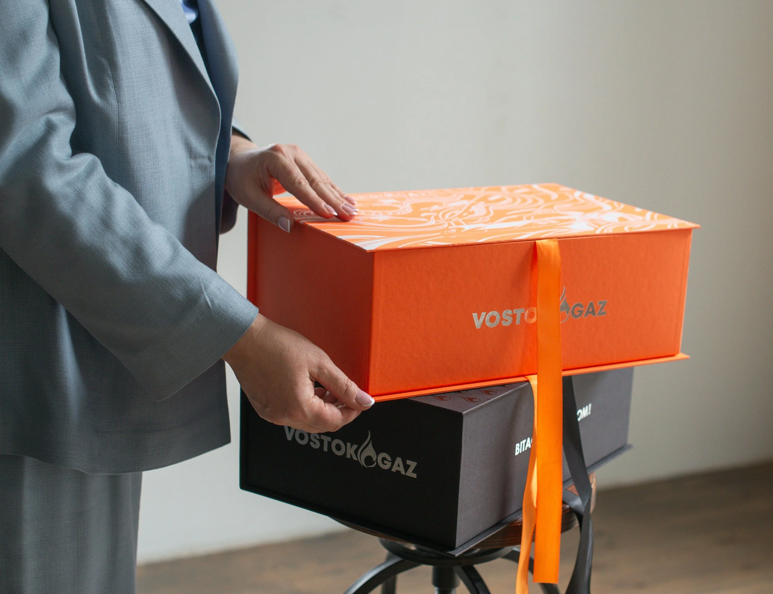 A person in a gray suit holding an orange box with a floral pattern on top and a ribbon. The box is labeled 'VOSTOK GAS.'