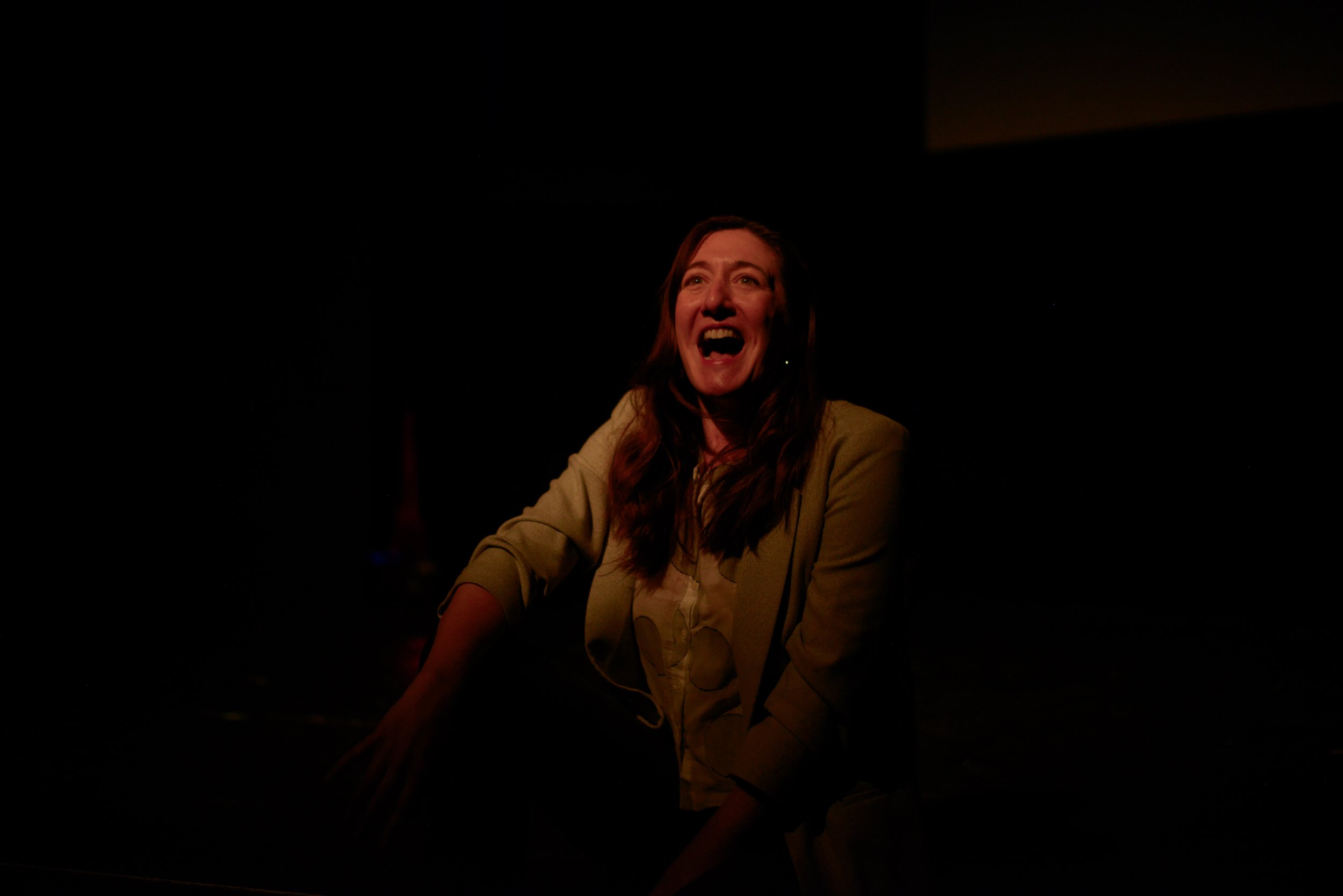 A woman with long brown hair and a light beige blazer laughing or speaking in a dark setting.