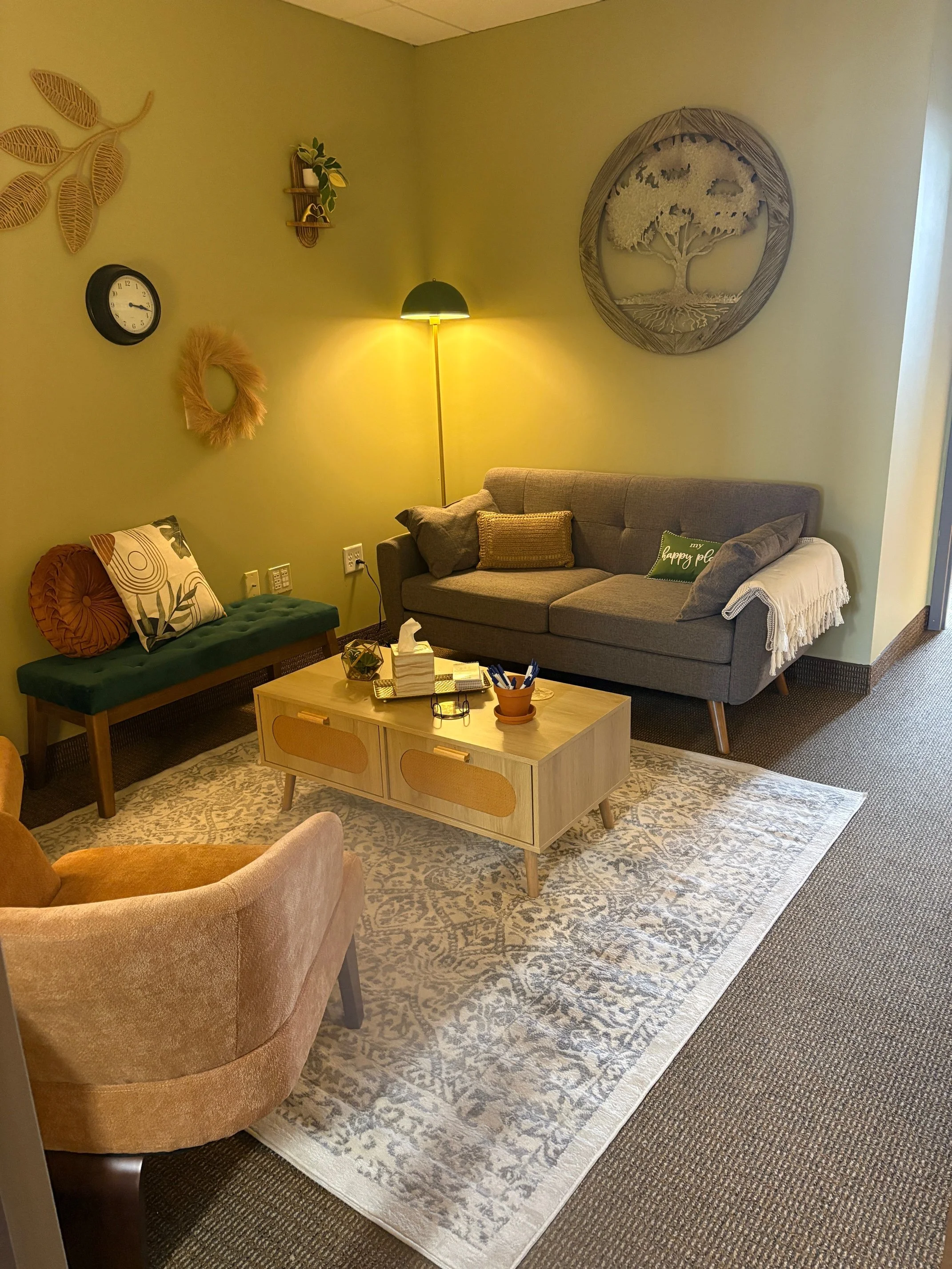 Cozy living room with a tan chair, green bench with pillows, gray sofa with cushions and blanket, wooden coffee table, patterned rug, wall decorations including a clock, wreath, art piece, small shelf with plant, and a floor lamp.