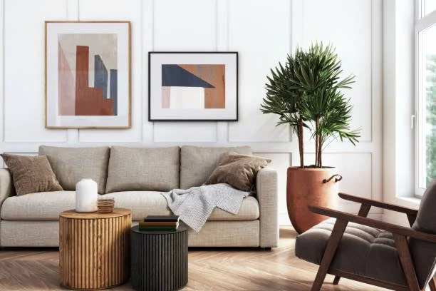 Inviting Living room with a beige sofa, abstract art prints on the wall, a large potted plant, and two round side tables, one wooden and one black, near a window.