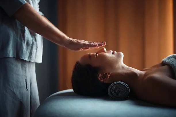 Person receiving Reiki, lying on a massage table with a rolled towel under their neck, while a therapist hovers their hands over their forehead gently.