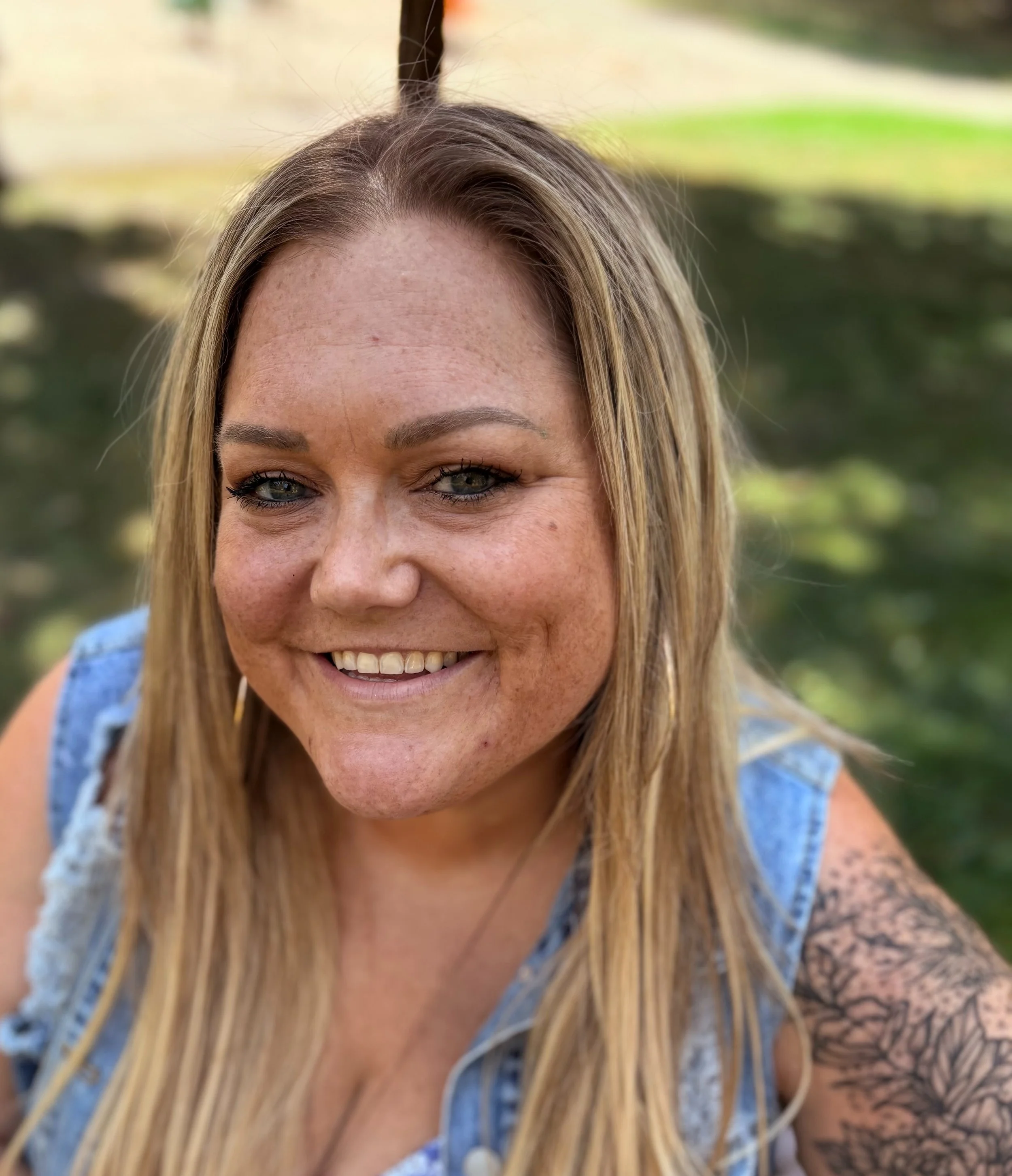 Close-up of a smiling woman with long blonde hair, wearing a denim vest, outdoors near a body of water with trees in the background.