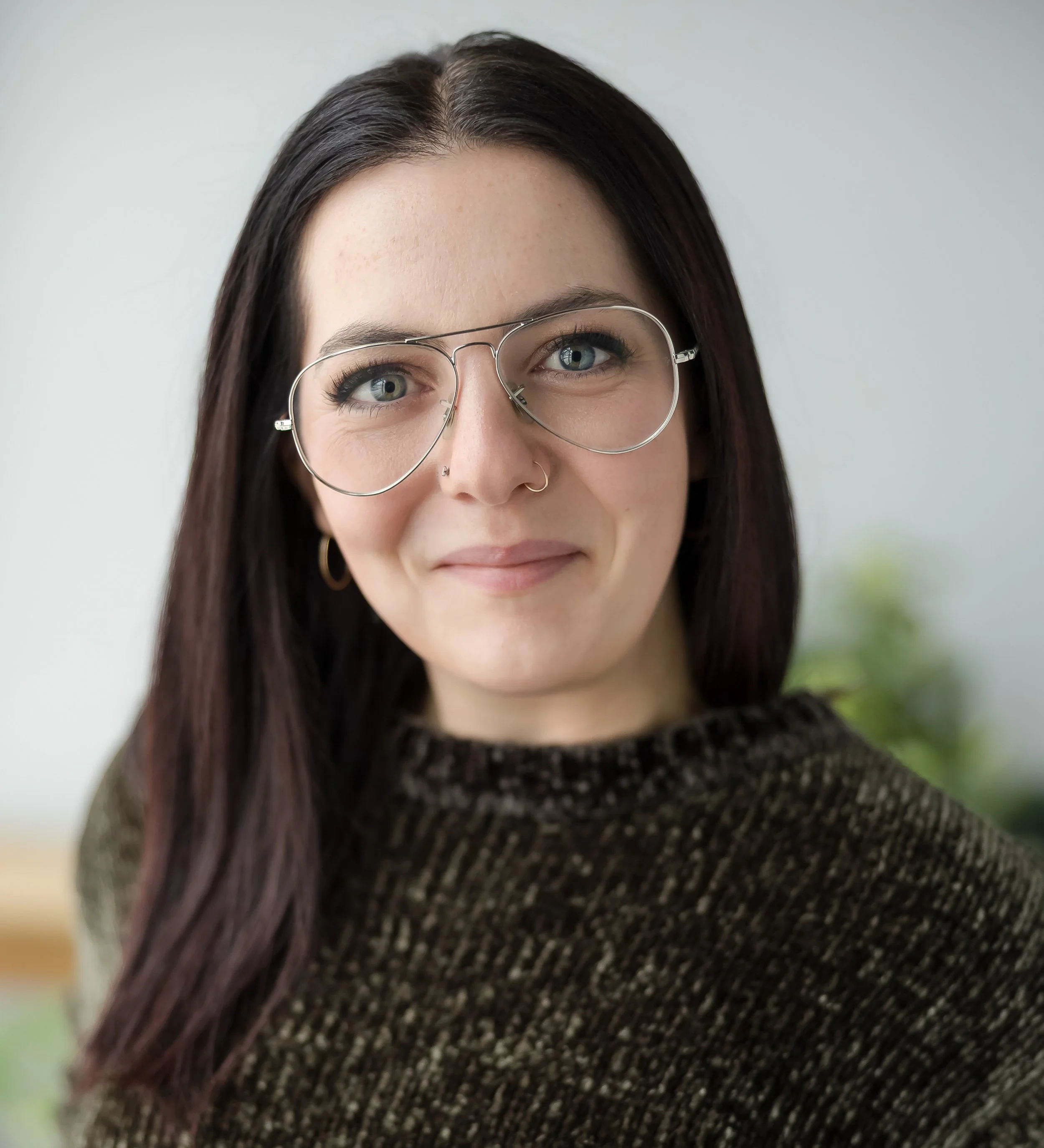 A woman with dark brown hair and blue eyes wearing clear, oversized glasses and a dark patterned sweater, smiling in front of a light-colored background with a blurred plant in the background.