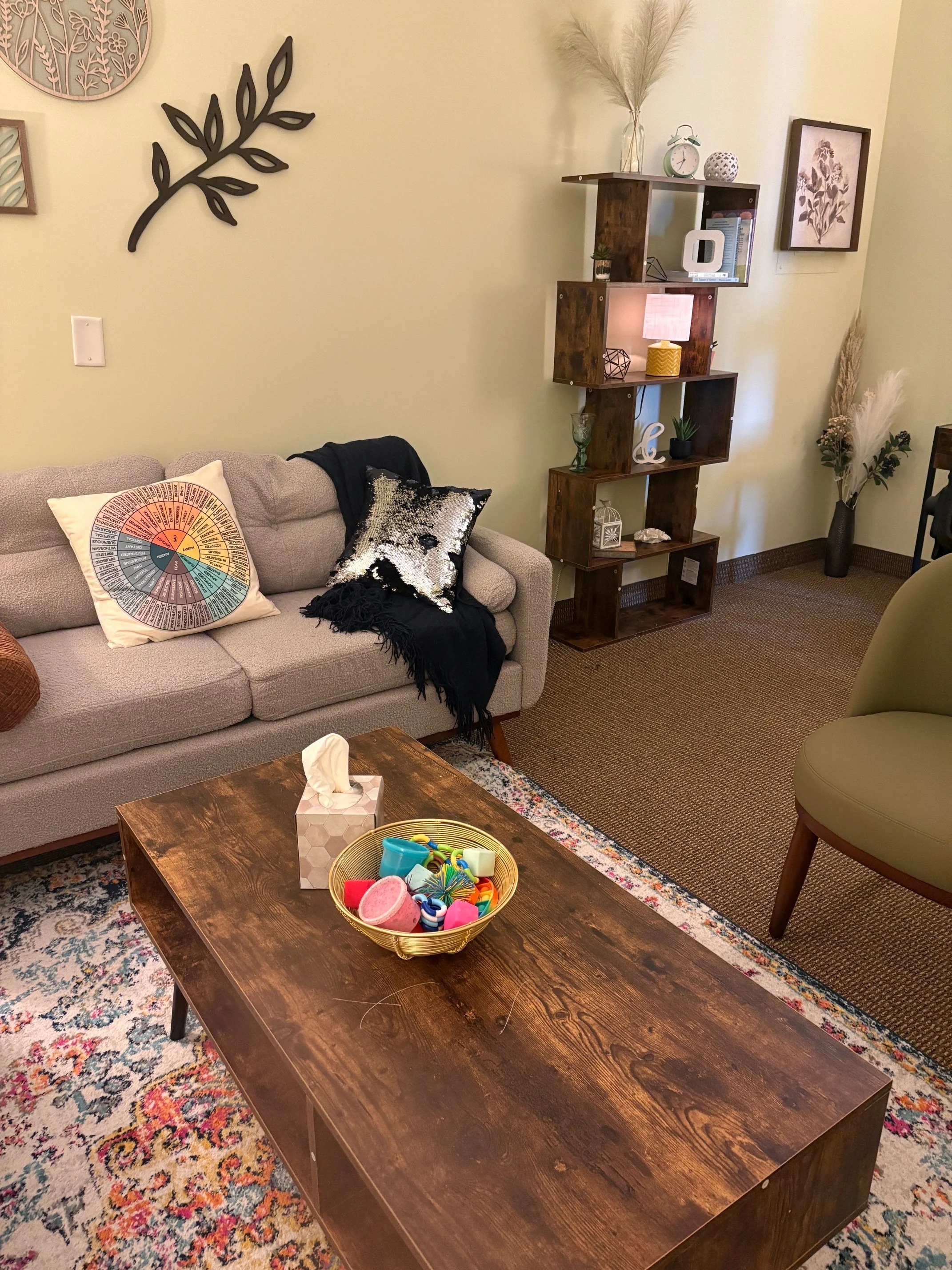 Living room with a beige sofa, decorative pillows, a wooden coffee table with toys, a patterned rug, a green armchair, and a wooden wall shelf with plants and decor. Artwork on the walls and dried flowers in vases are also visible.