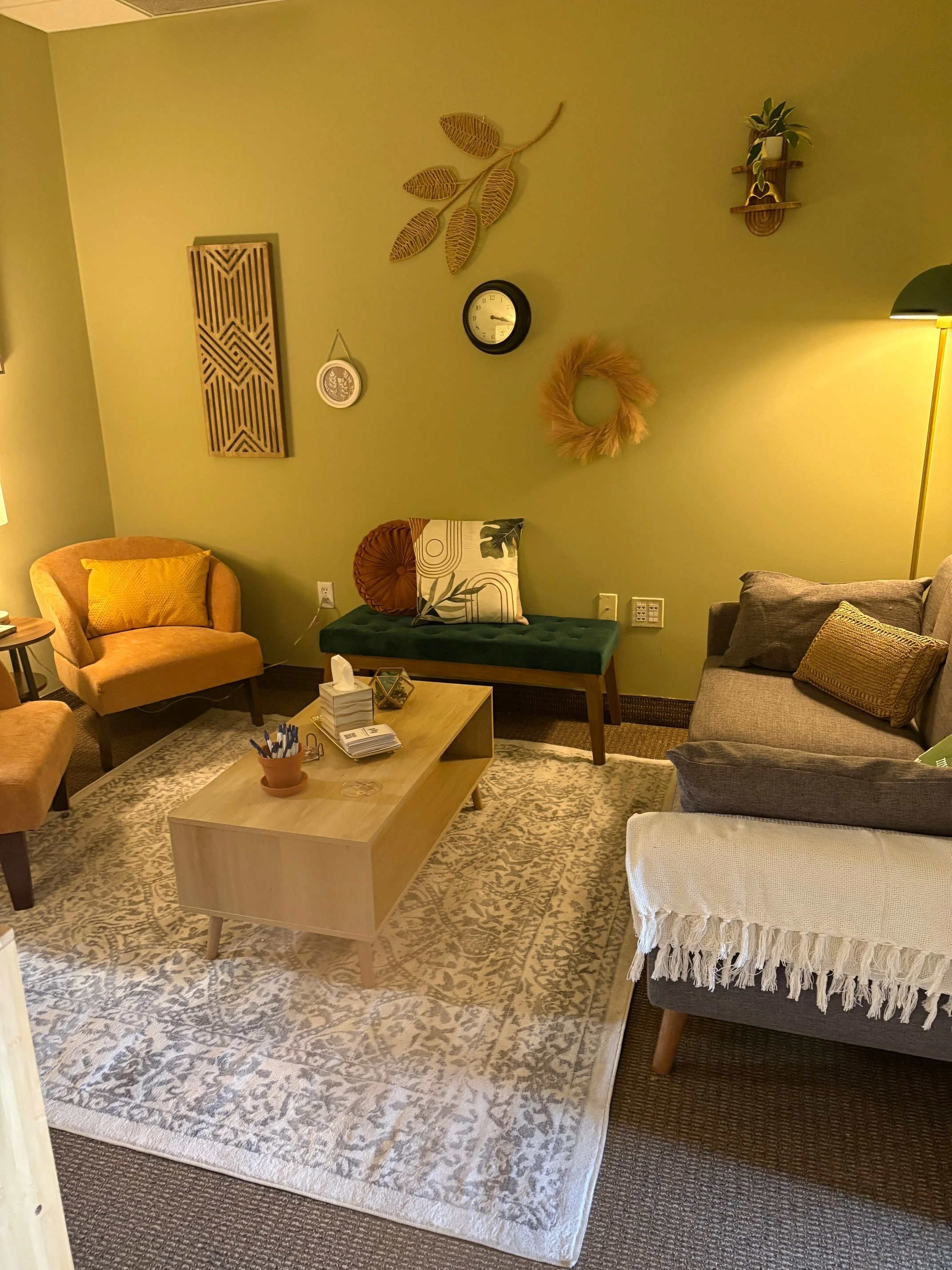 Cozy living room with beige, gray, and green furniture, including a green bench, a gray sofa with pillows, and two yellow armchairs, with a wooden coffee table on a patterned rug. Wall decor includes a clock, art pieces, a wreath, and a small shelf w