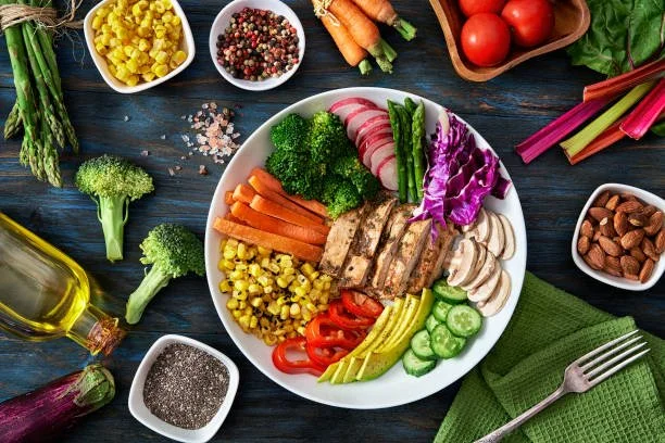 A beautiful plate of colorful sliced vegetables and chicken. Surrounding bowls contain cherry tomatoes, almonds, mixed peppercorns, and chopped vegetables on a dark wooden table.