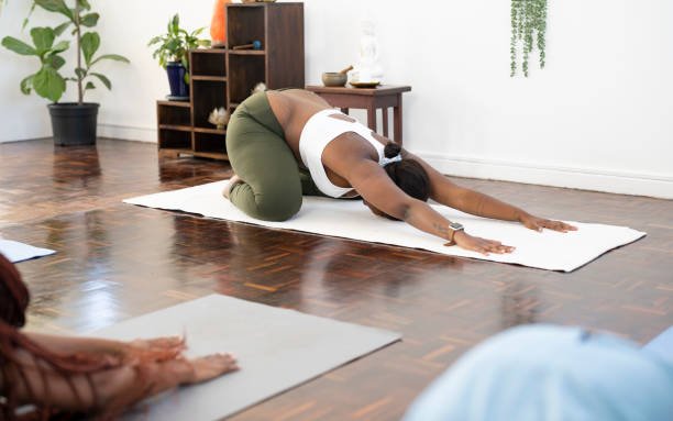 Woman practicing yoga in child's pose on a white mat in a home studio with wooden floor and plants.