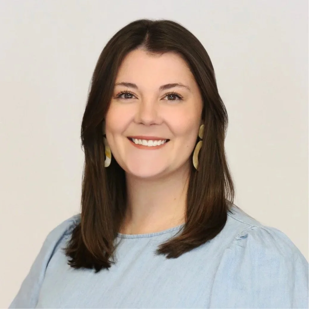 Portrait of a woman with shoulder-length brown hair, smiling, wearing a light blue top and yellow earrings, against a plain light background.