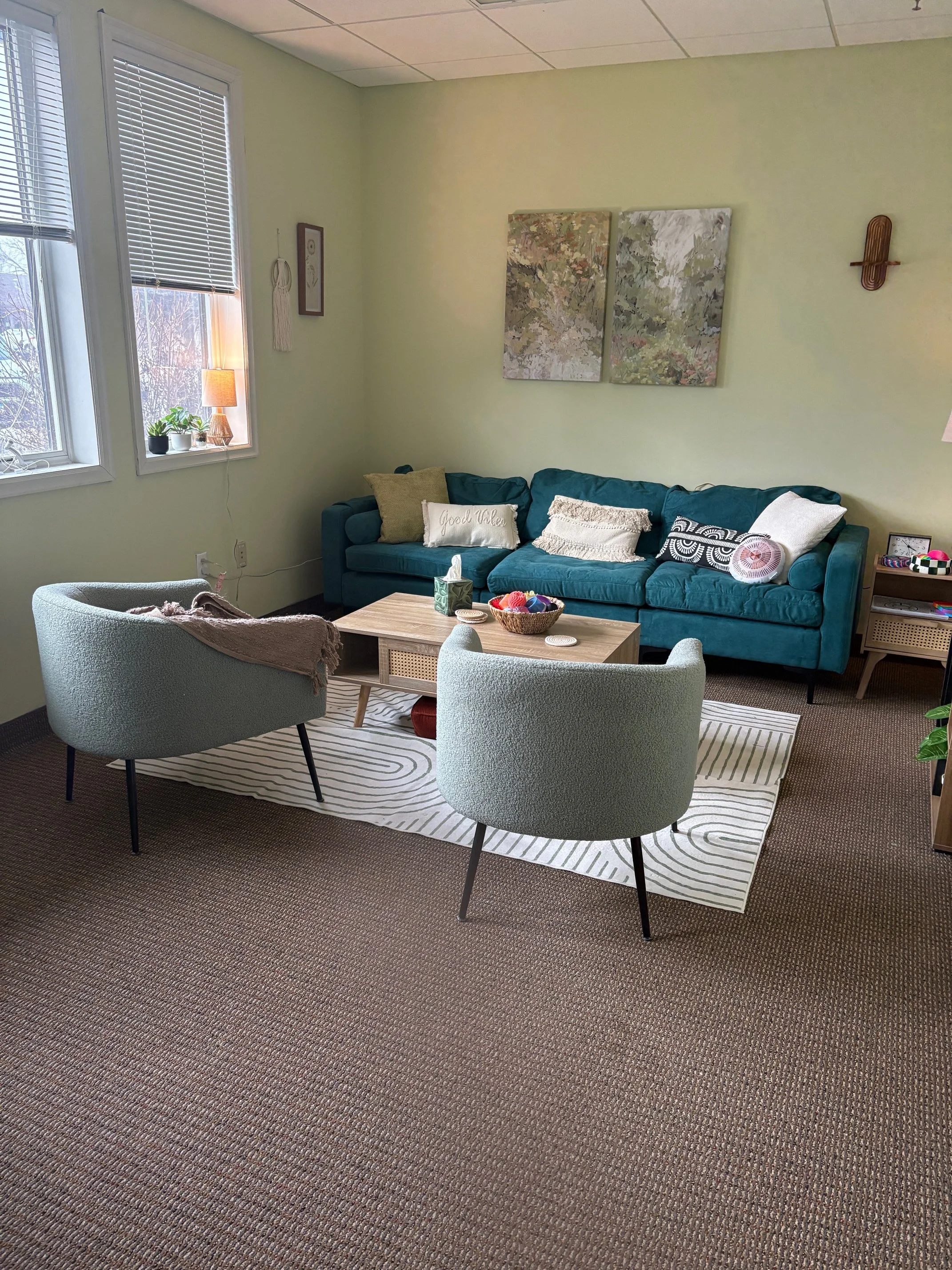 A cozy living room with a teal sofa, two light-colored armchairs, a wooden coffee table, a rug, a window with blinds, and decorative wall art.