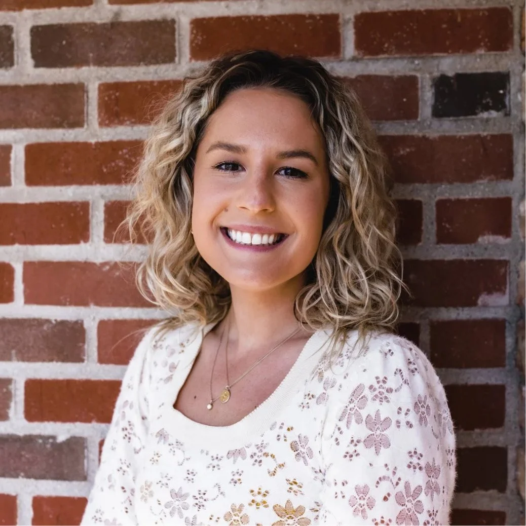 A young woman with curly blonde hair smiling, wearing a white floral lace top and standing in front of a brick wall.