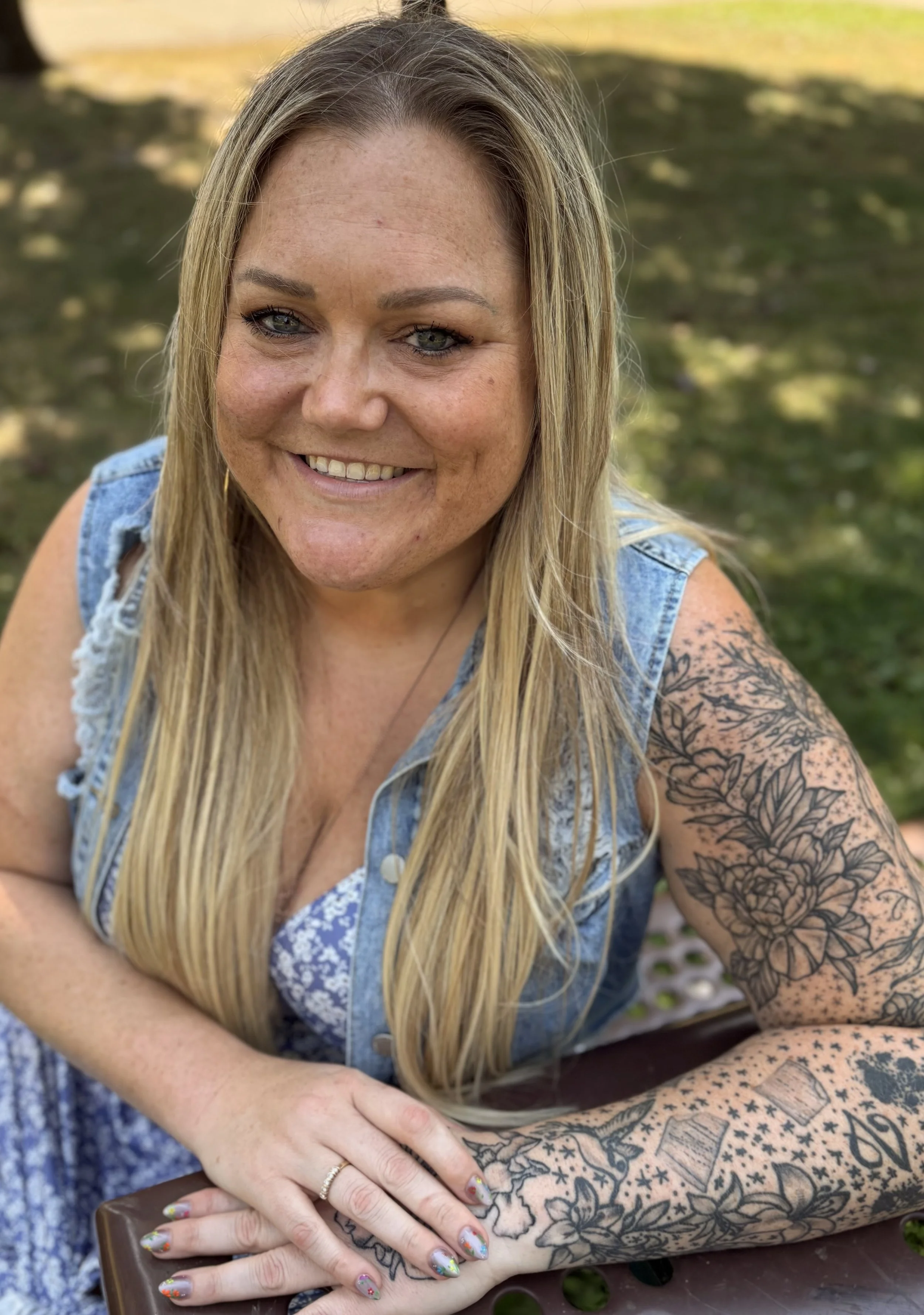 Smiling woman with long blonde hair and tattoos on her right arm, wearing a sleeveless denim vest and sitting outdoors with green grass in the background.