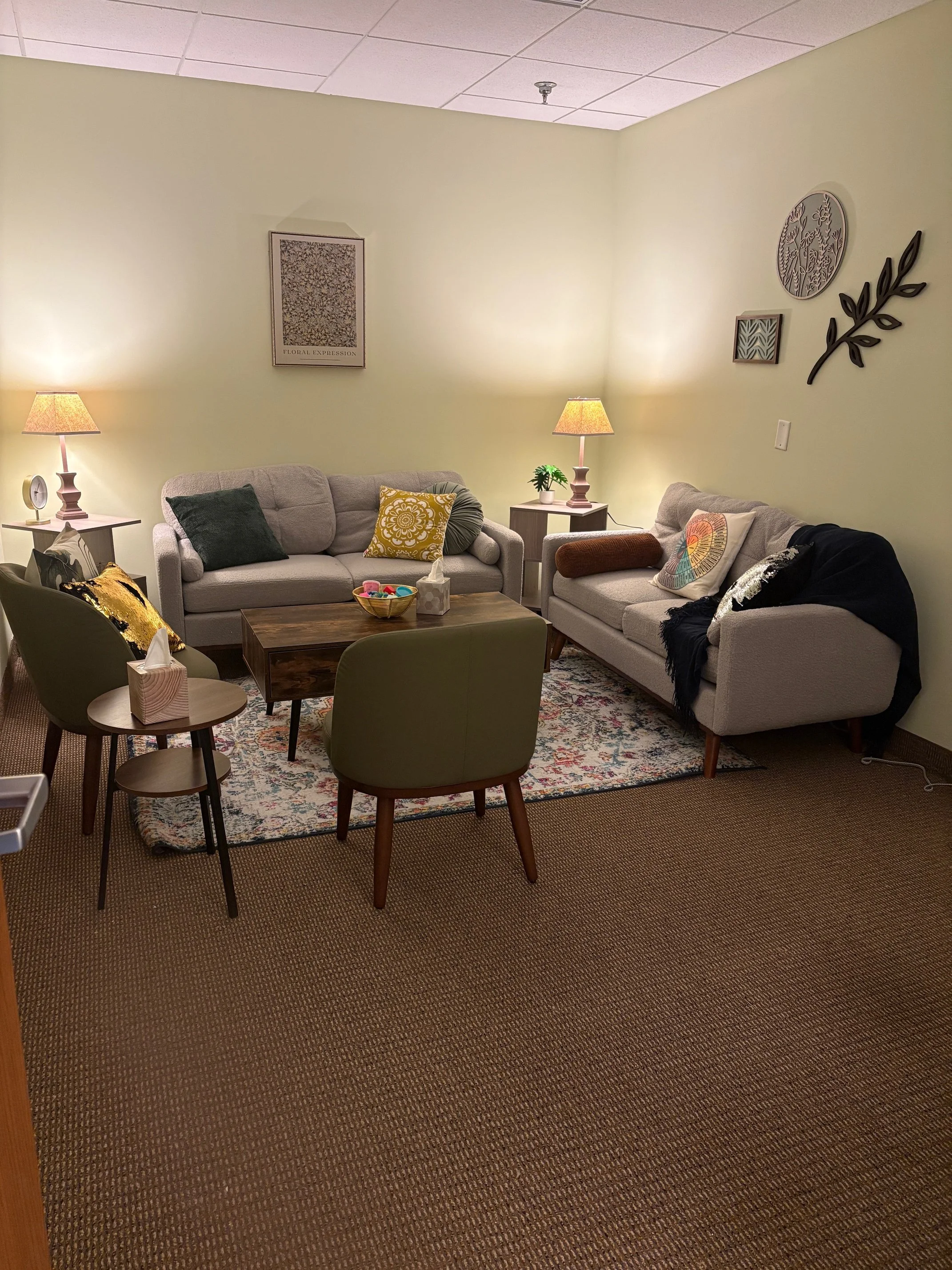 A cozy living room with two light-colored sofas, a dark wood coffee table, and three side tables with lamps. The couches have colorful pillows and throws. There is a patterned rug under the coffee table, and wall decorations including framed art and 