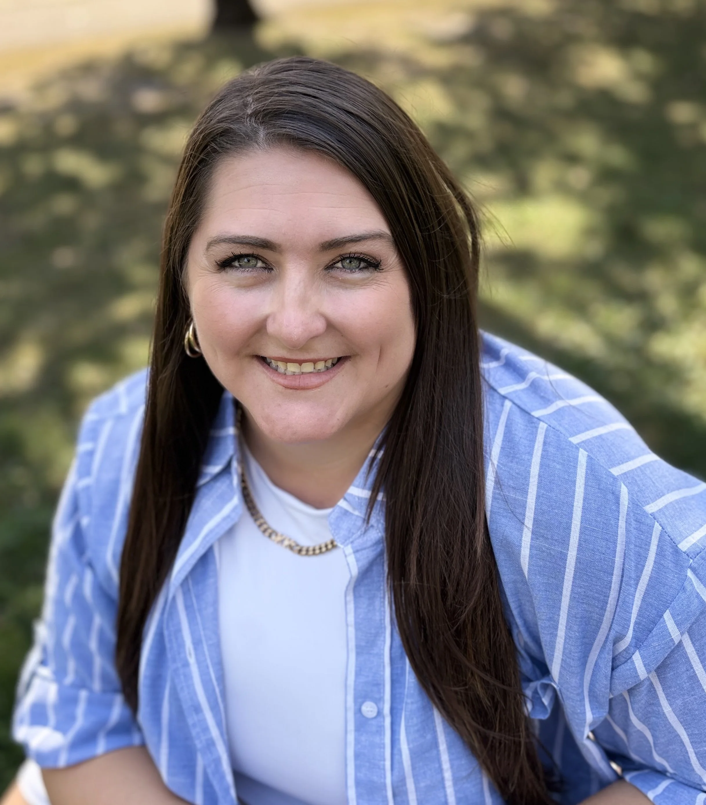 Close-up portrait of a woman with long brown hair, blue eyes, and a friendly smile, wearing a blue and white striped shirt and gold jewelry, outdoors with greenery in the background.