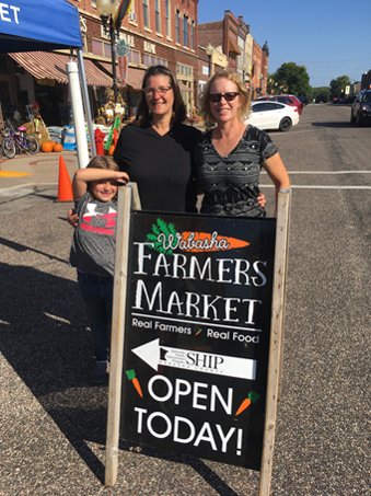 Sara George and daughter, Faith (left). Tina Moen (Right) standing by farmers market sign saying open today.png