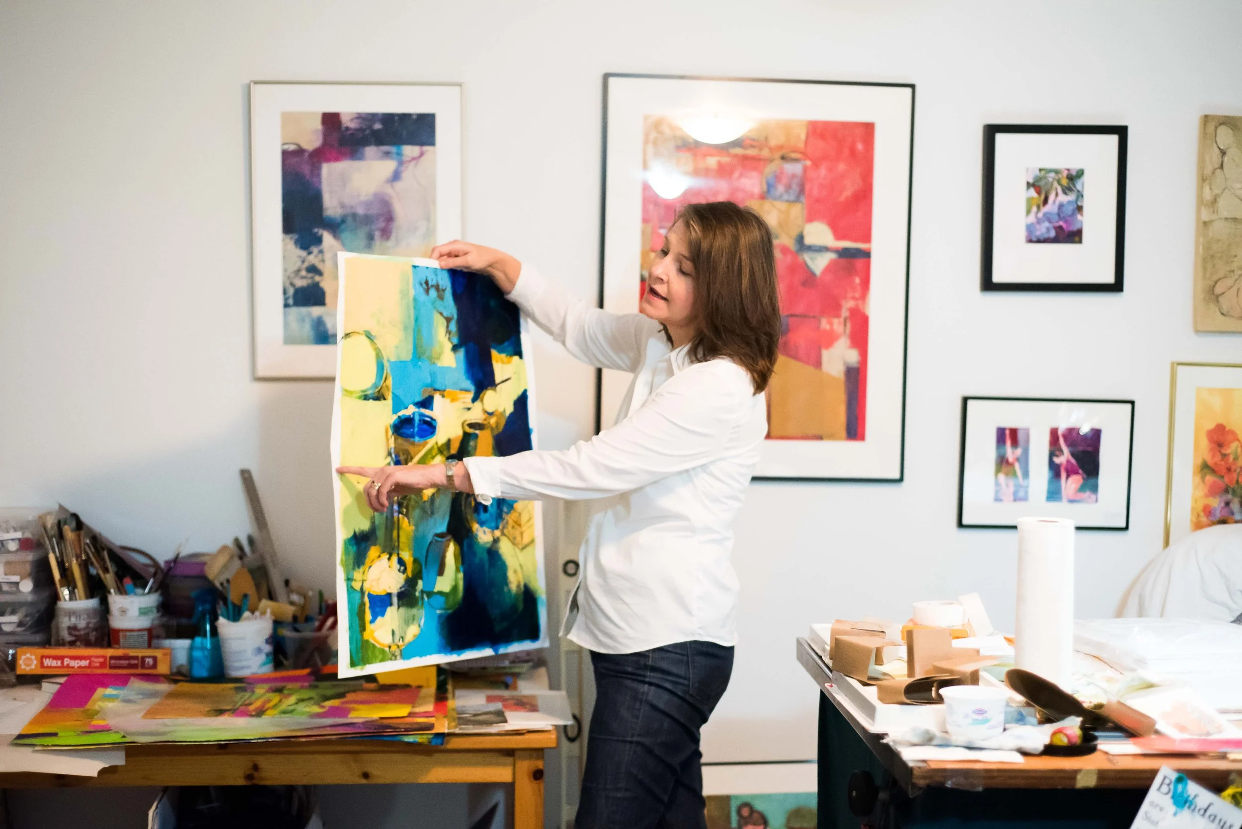 Carolyn holds up a painting in process in her studio space surrounded by paintings and painting supplies