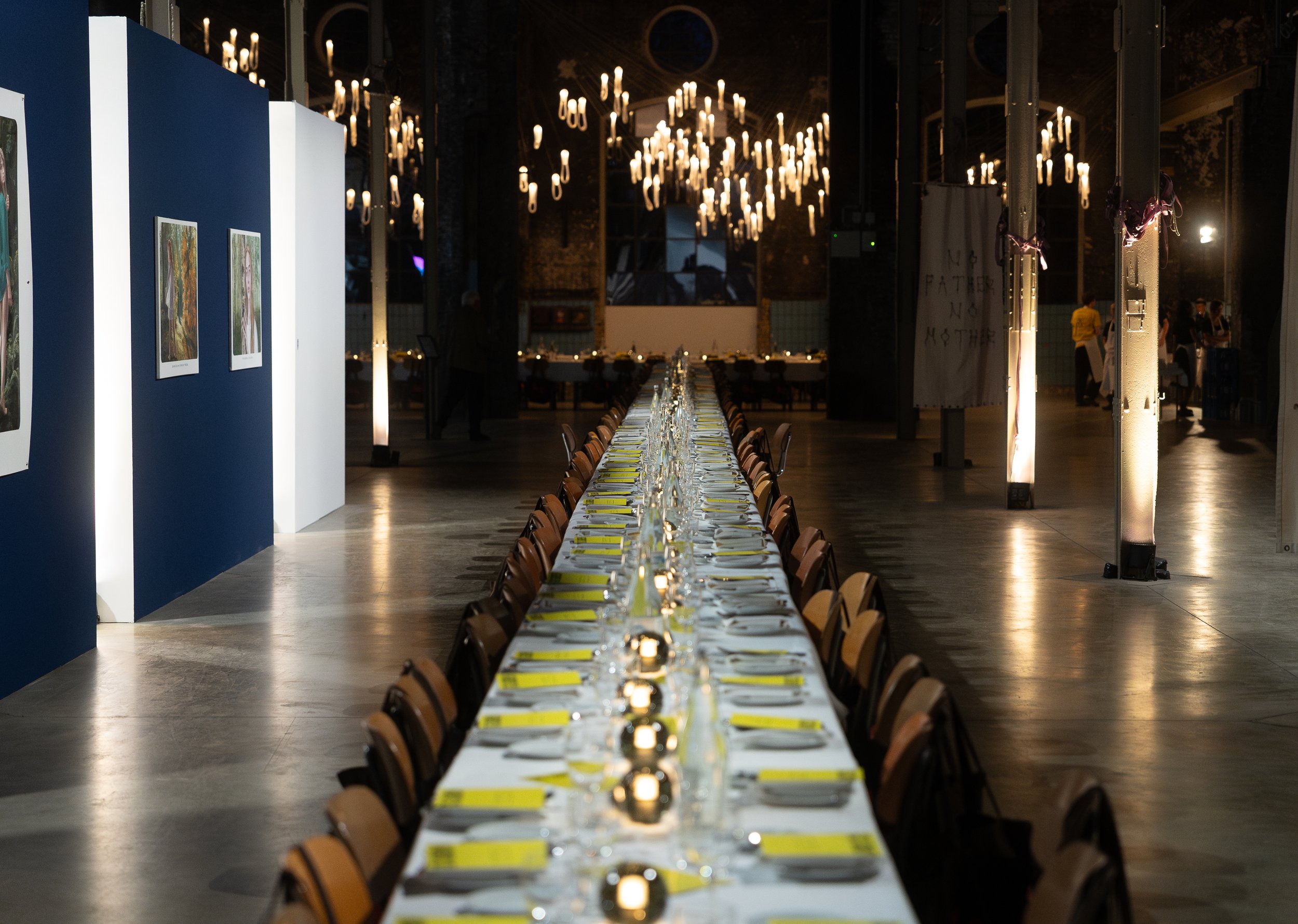 Langer, festlich gedeckter Tisch in einem großen Raum mit Kerzenleuchtern und Wanddekorationen; links Gemälde an einer blauen Wand.