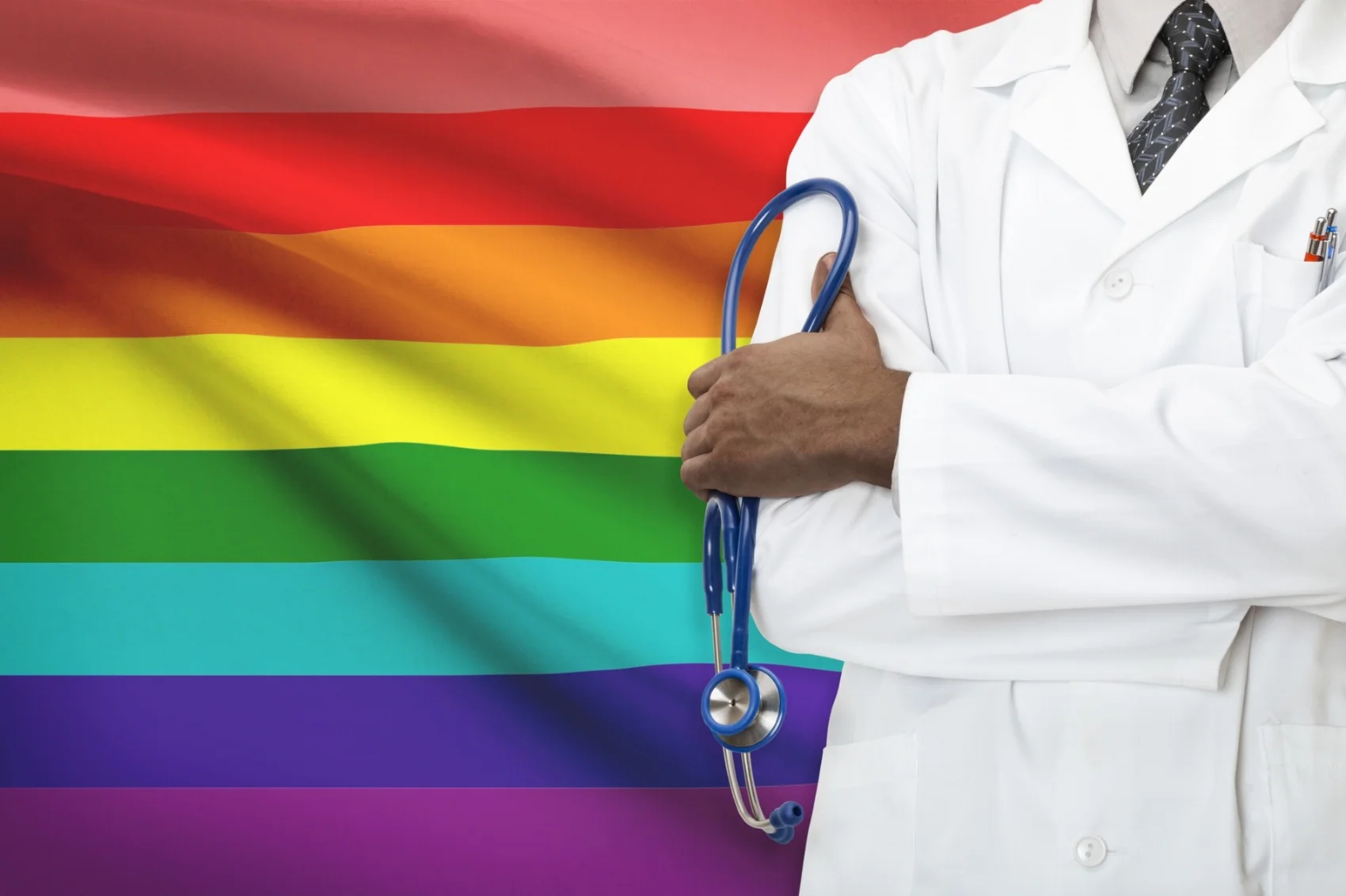 Medical professional (from the shoulders down) with stethoscope standing in hand arms crossed standing against a Pride flag. 
