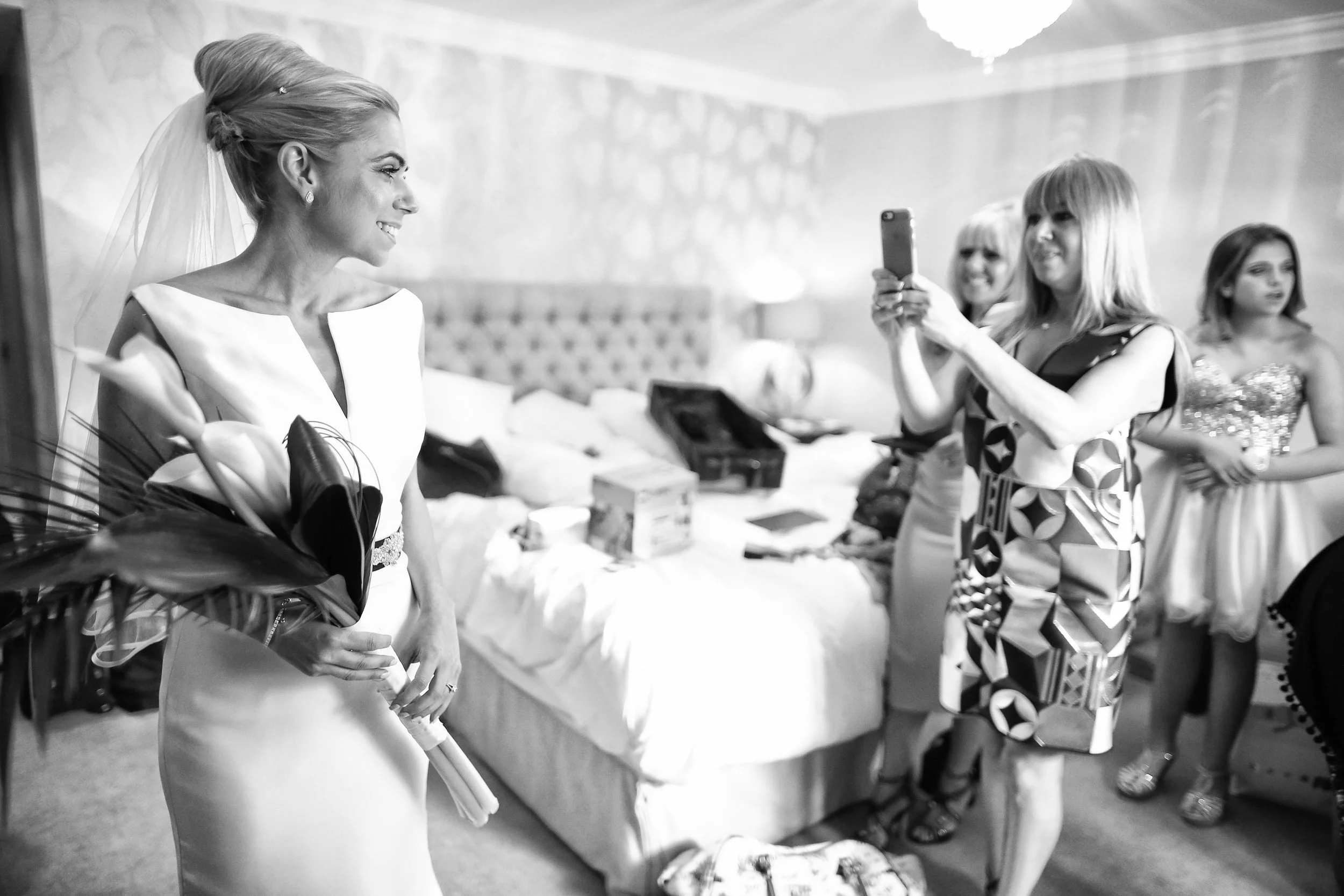 Bride holding bouquet, being photographed by women in a bedroom setting.