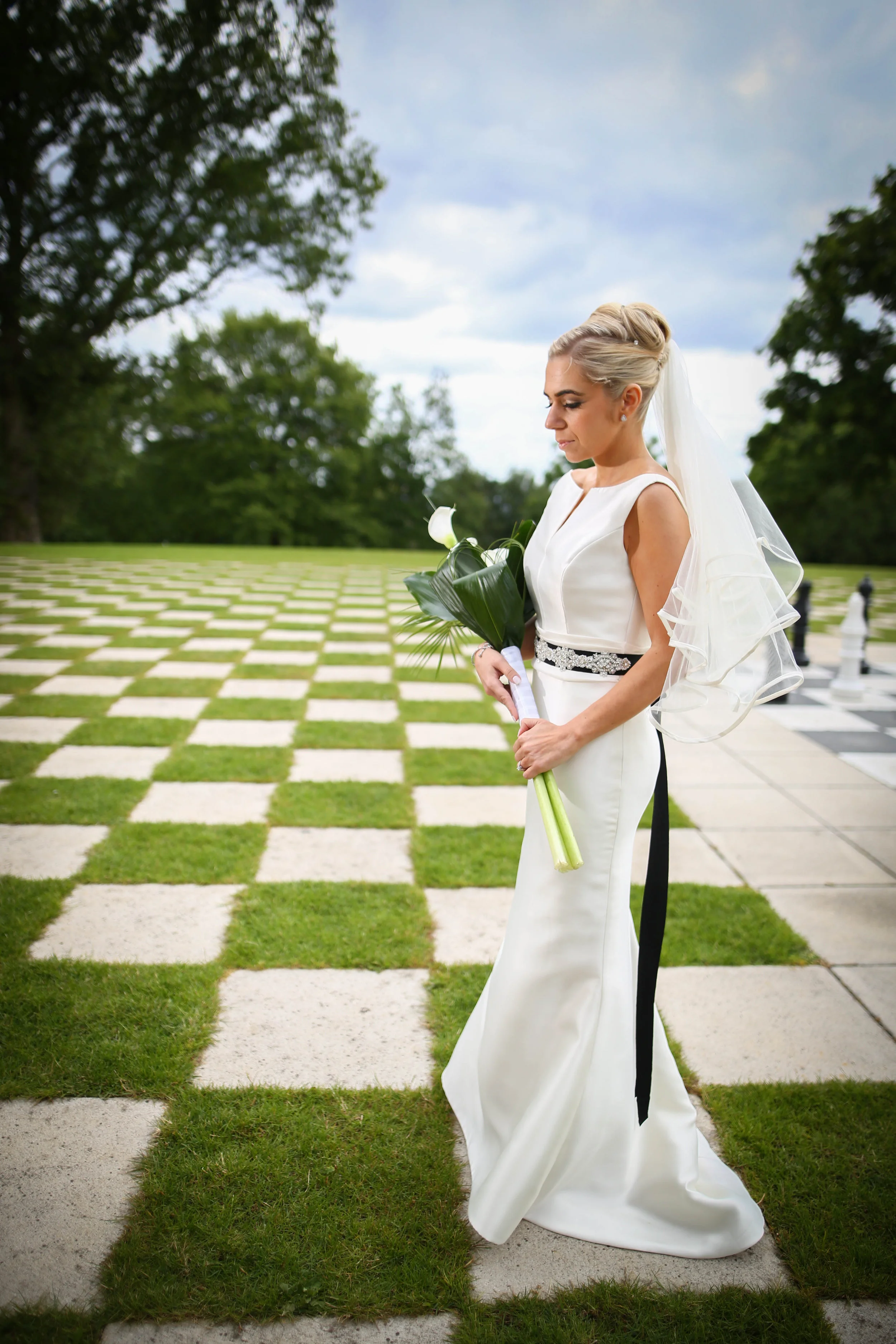 Bride in white dress with black sash holding bouquet of calla lilies, standing on checkered stone and grass path in garden setting.