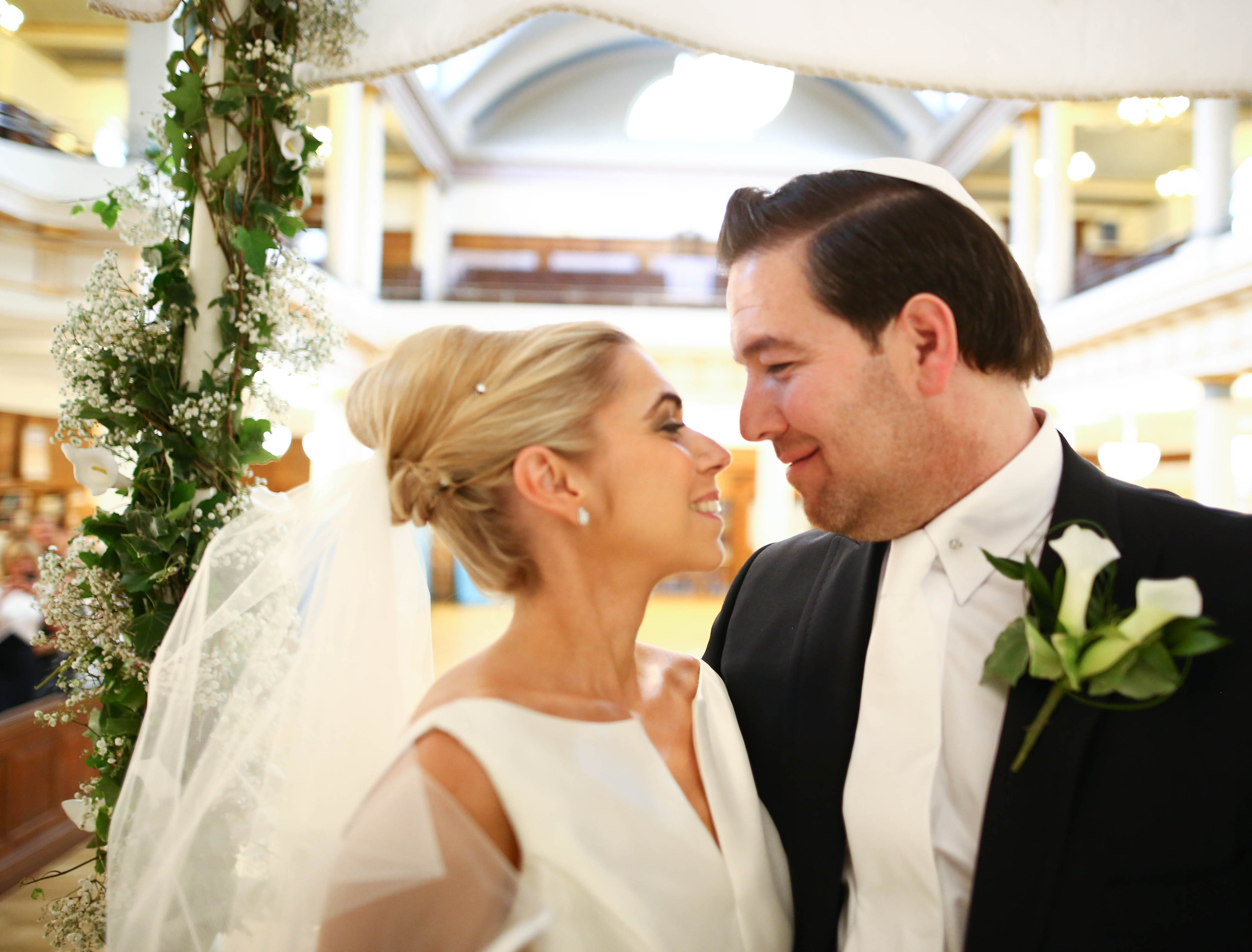 Wedding couple under chuppah, bride in white dress with veil, groom in black suit with boutonniere, standing in synagogue.