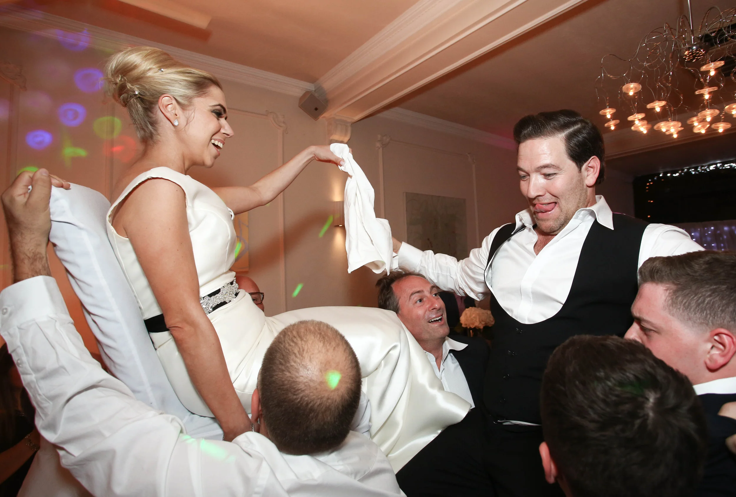 A wedding celebration scene where a bride and groom are lifted on chairs by guests, holding hands with a cloth between them, inside a decorated room with lights.