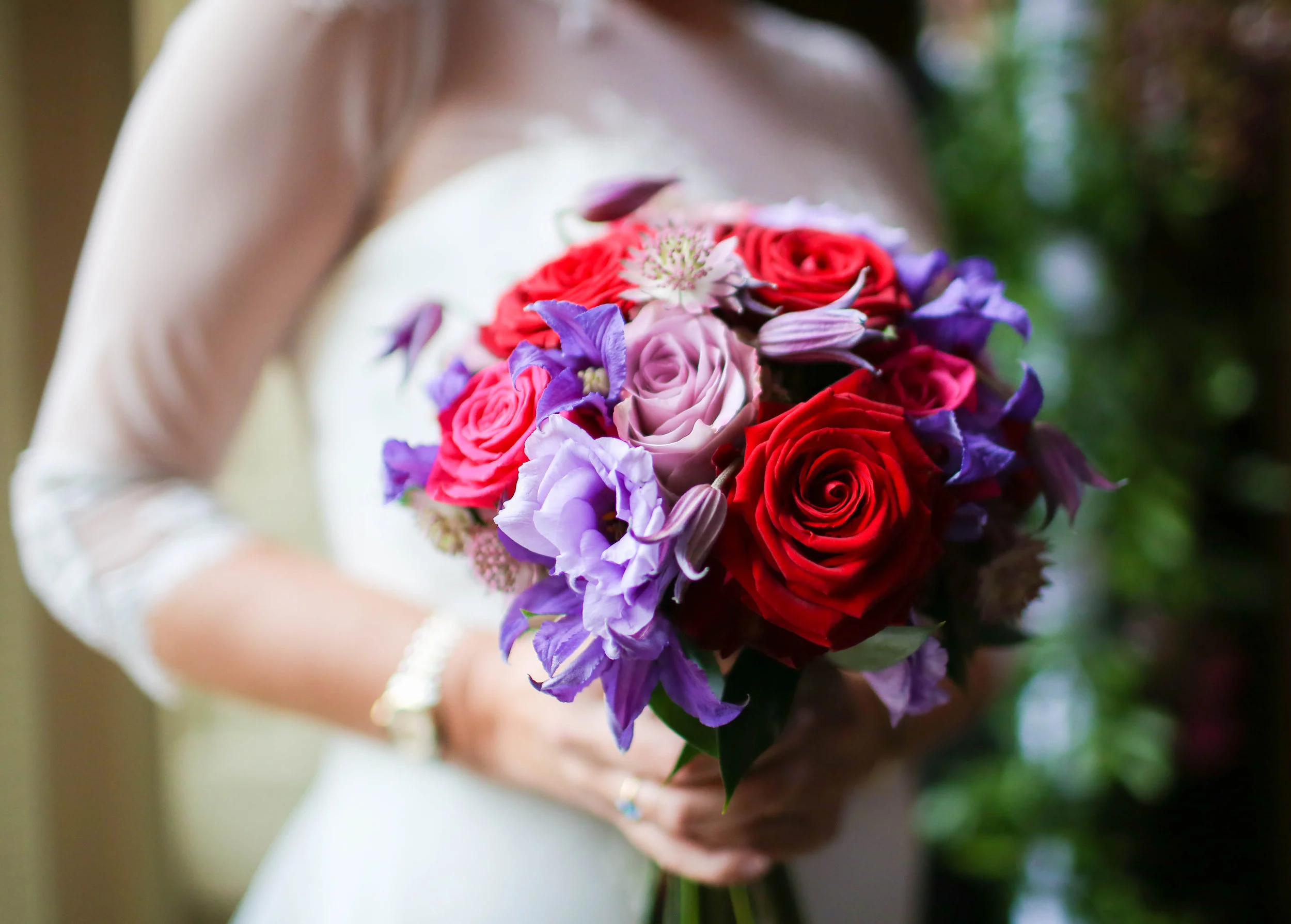 Bride holding a bouquet with red roses, purple flowers, and pink blooms.