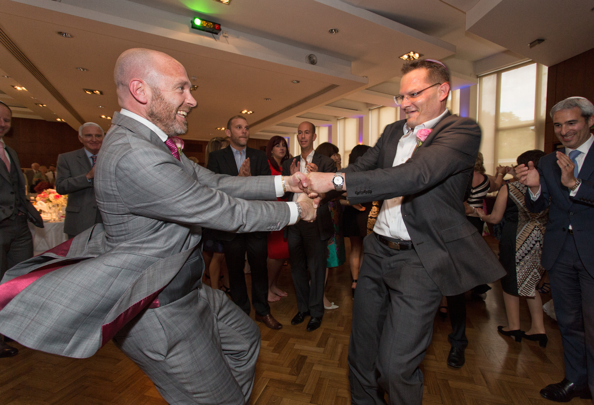 Two men in suits dancing joyfully at a wedding reception, surrounded by clapping guests.