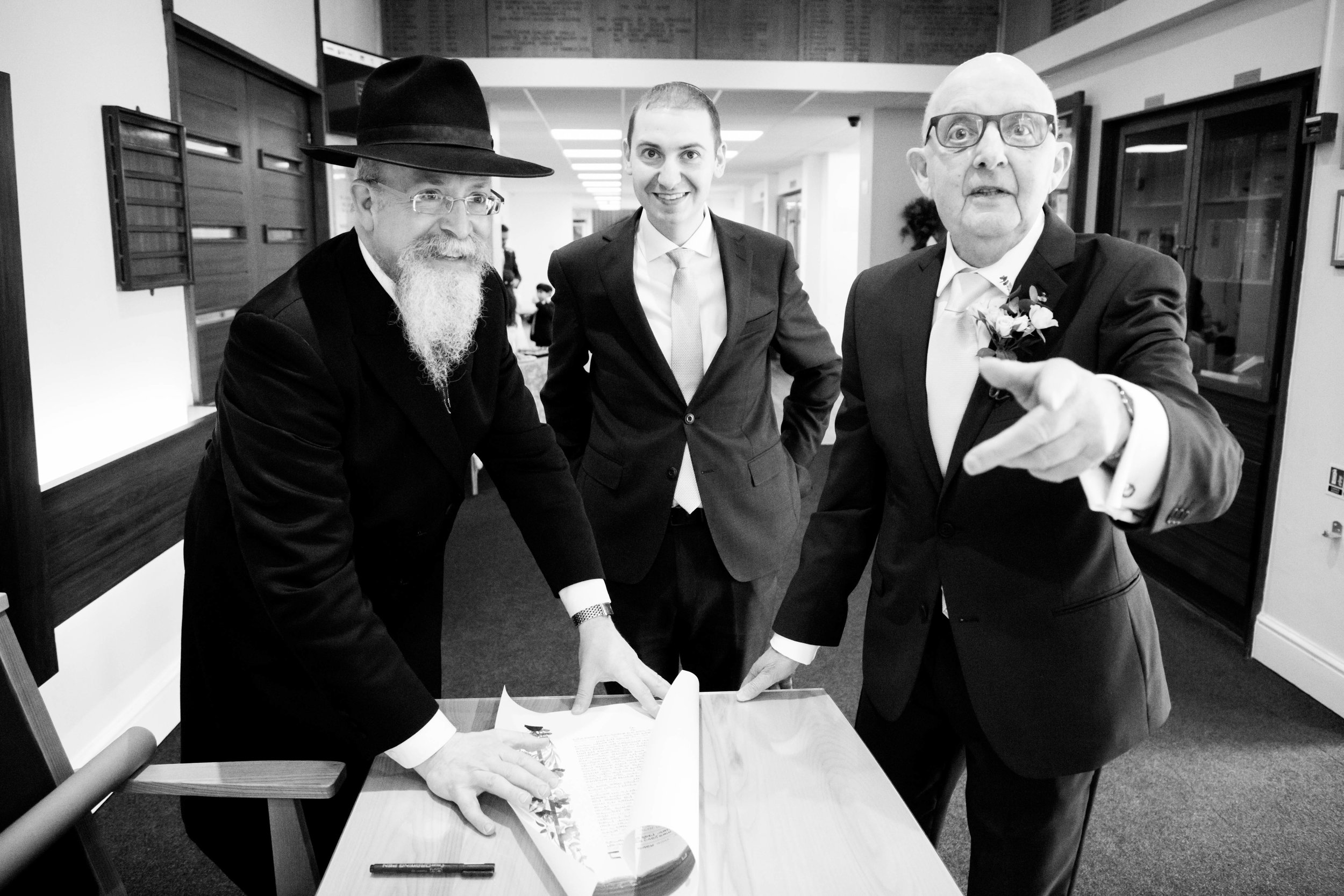 Three men in formal suits gathered around a table with a document, in a hallway. One man points forward, others look on smiling.