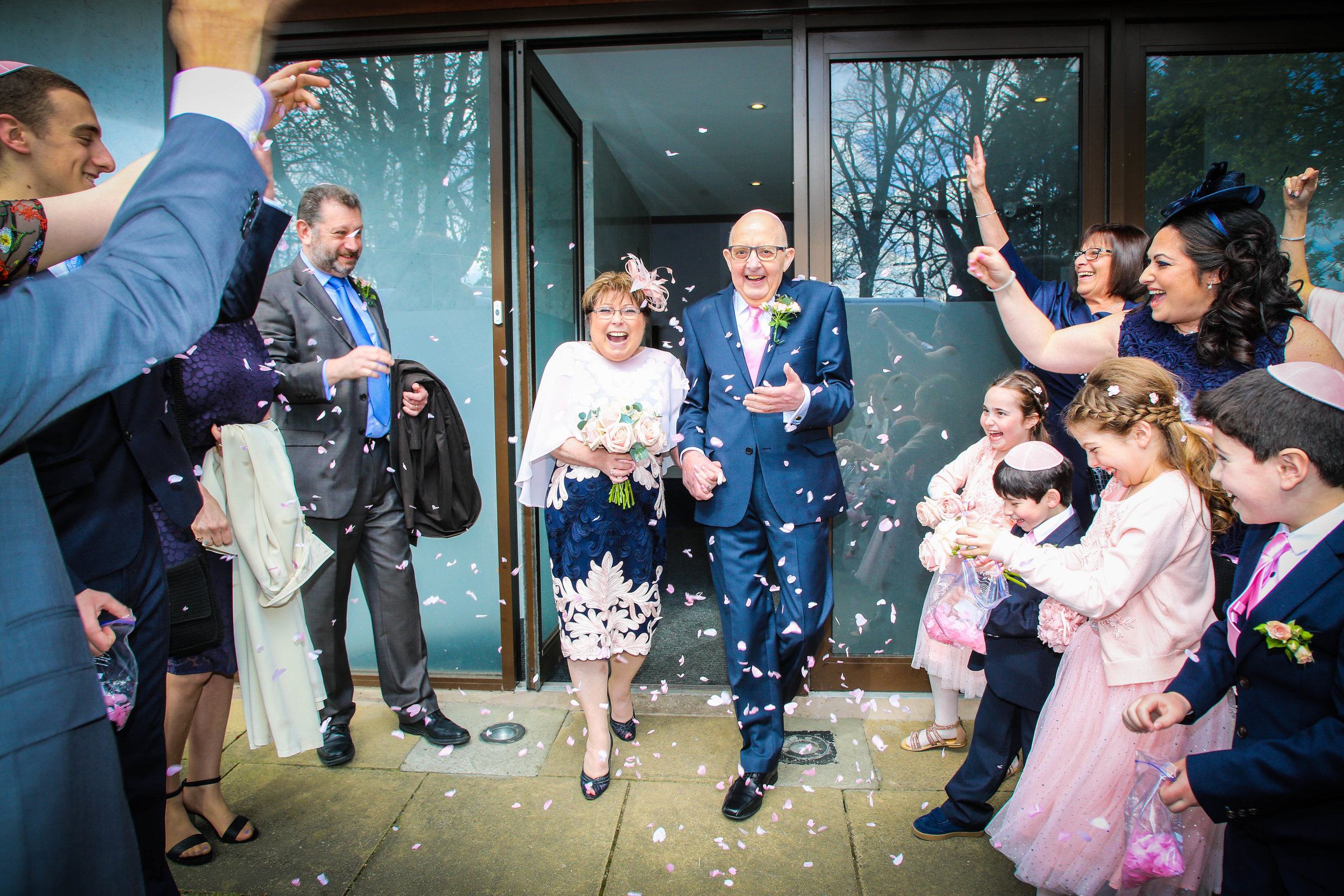 A joyful wedding exit with a bride and groom, dressed in formal attire, surrounded by smiling guests throwing flower petals.