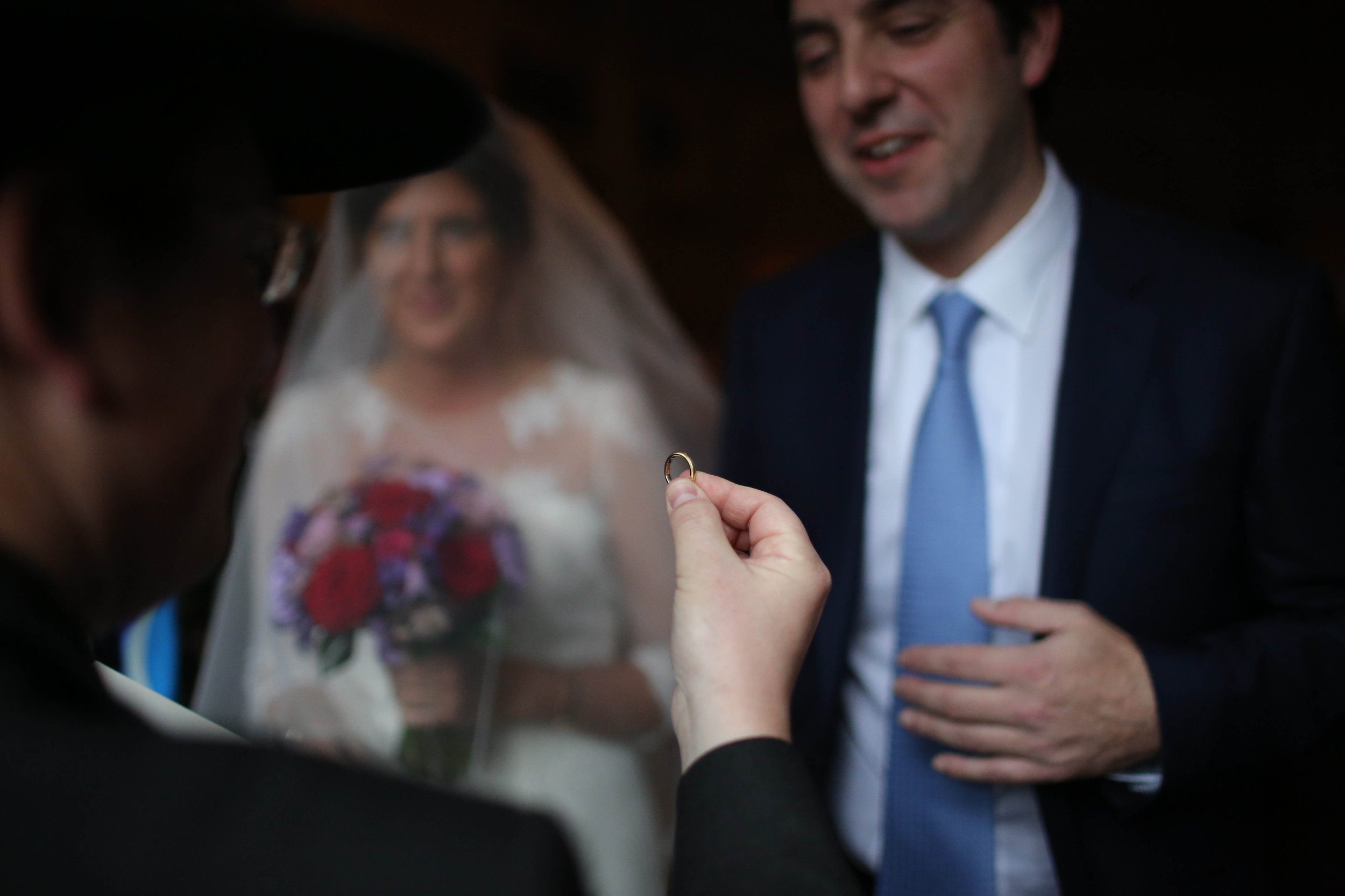 Close-up of a person holding a gold wedding ring, with a bride and groom in the background, during a wedding ceremony.