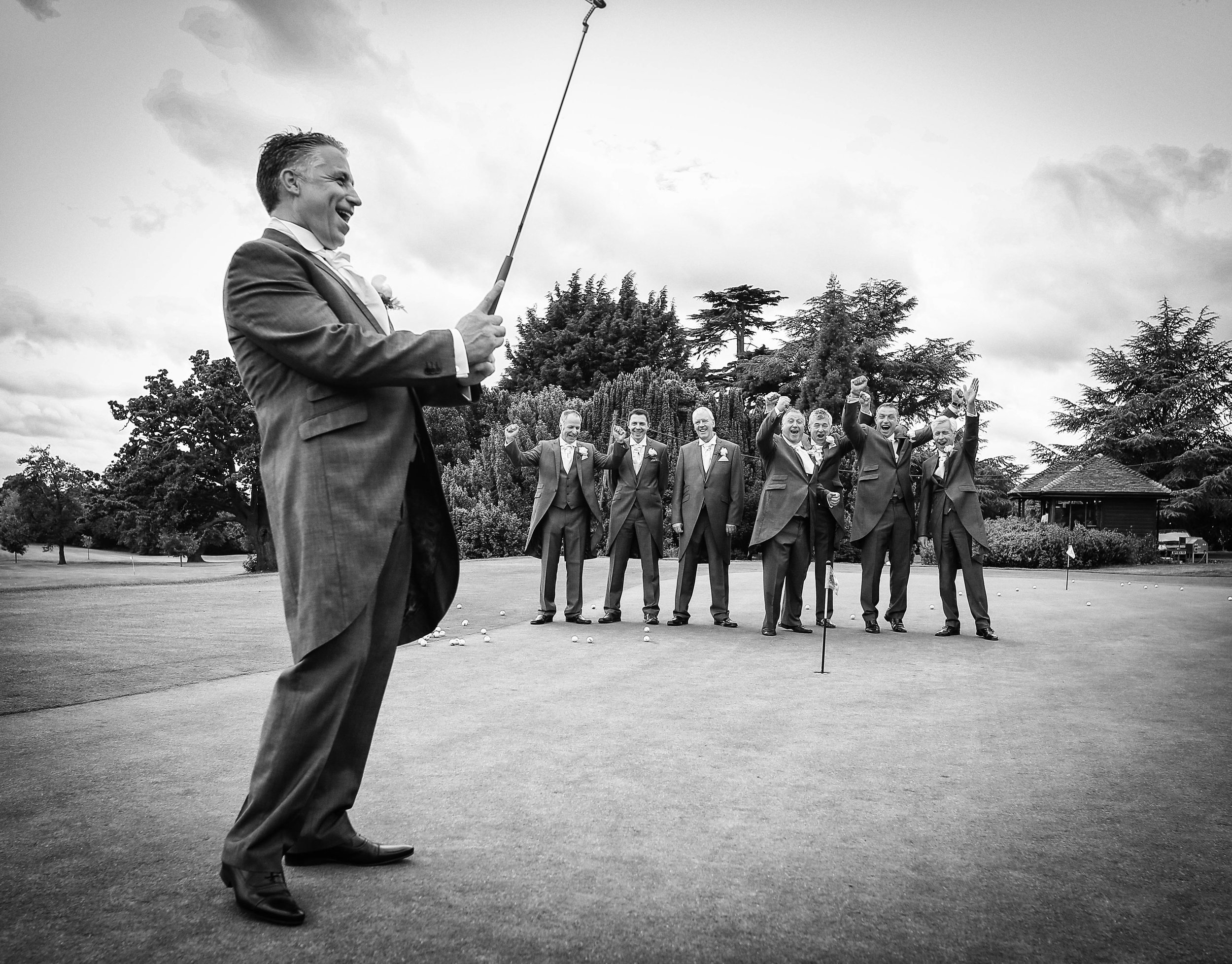 Group of men in suits on a golf course, one playing with a golf club, celebrating with raised hands.