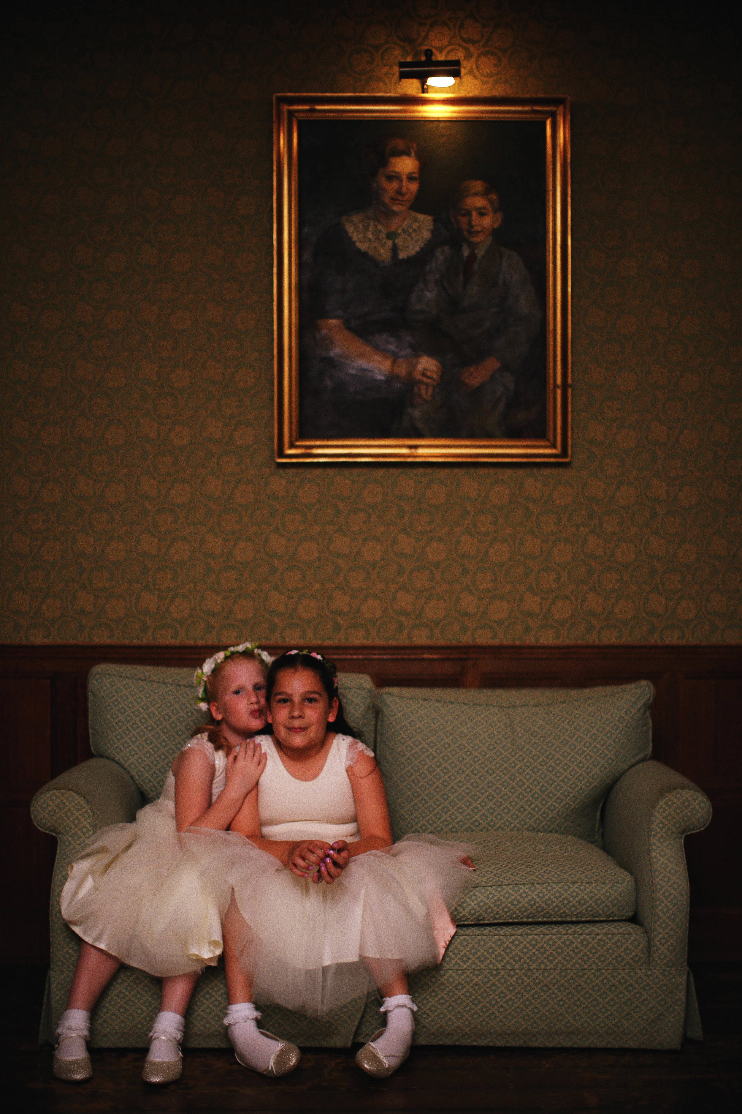 Two young girls in white dresses sitting on a green sofa with a portrait of a woman and a child hanging above them on the wall.