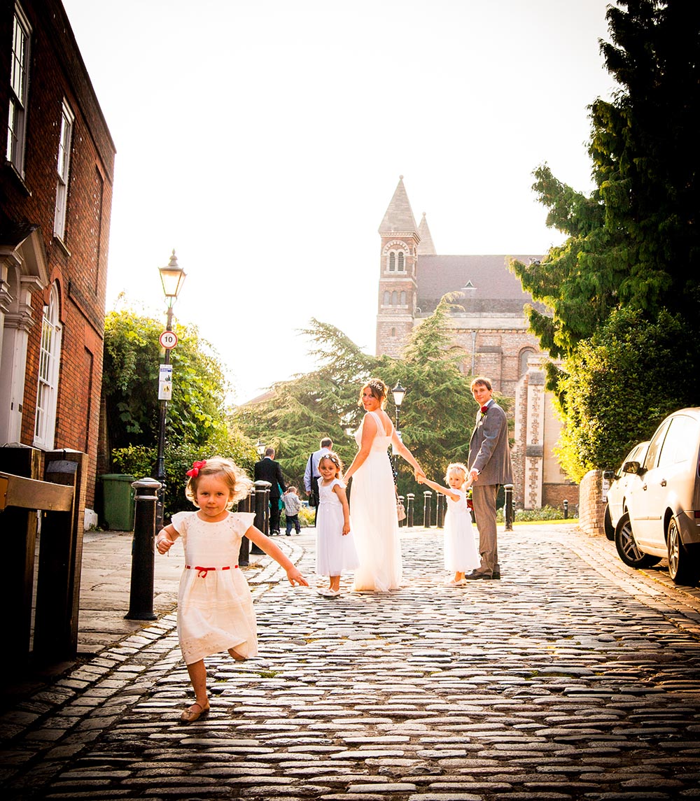 A wedding scene on a cobblestone street with a bride, groom, and children in formal attire, with a church and trees in the background.