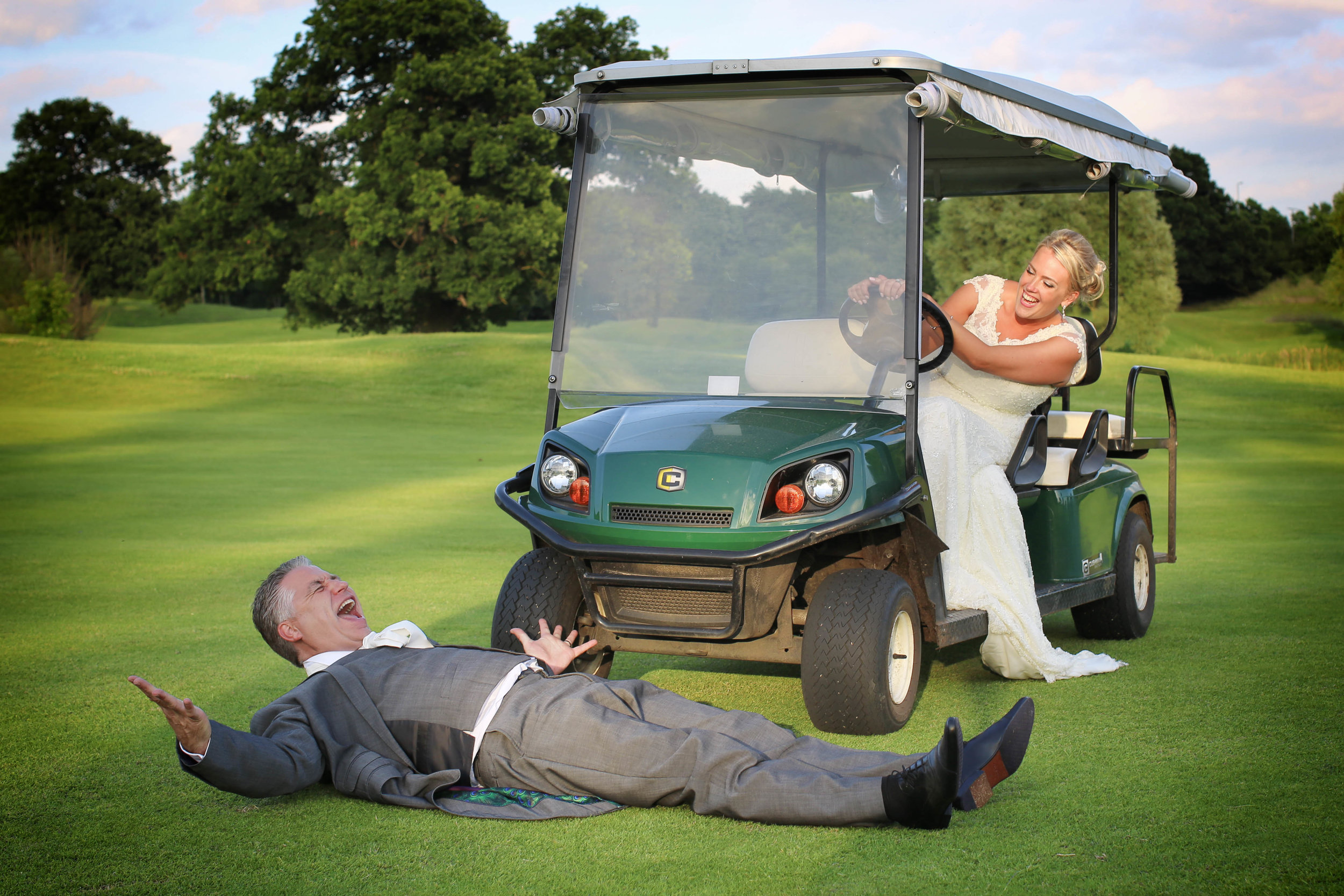 Bride driving a golf cart, groom playfully laying on the ground in front on a golf course.