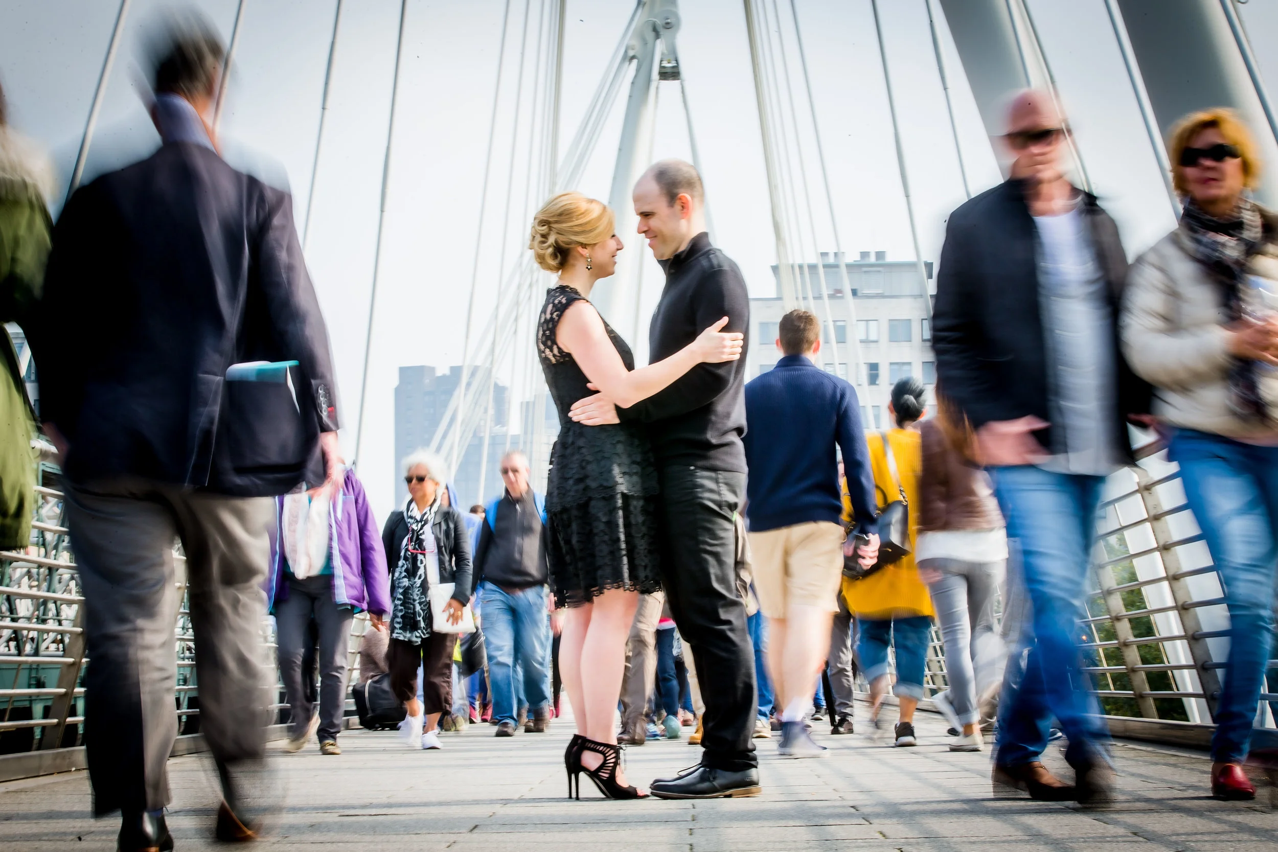Couple embracing on a busy pedestrian bridge, surrounded by blurred motion of people walking by.