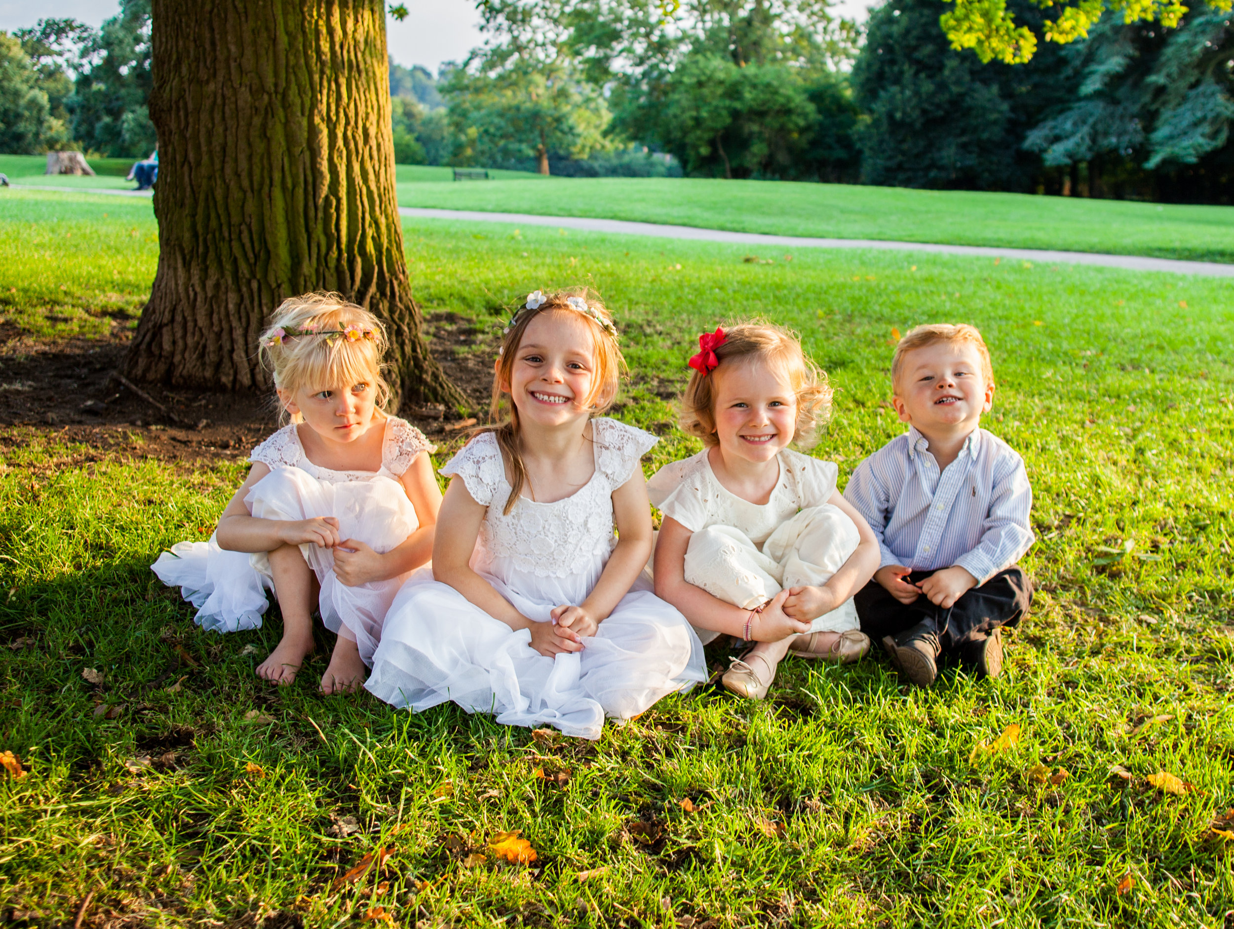 Four children sitting on grass by a tree in a park, three girls in white dresses and one boy in a white shirt, smiling and looking at the camera.