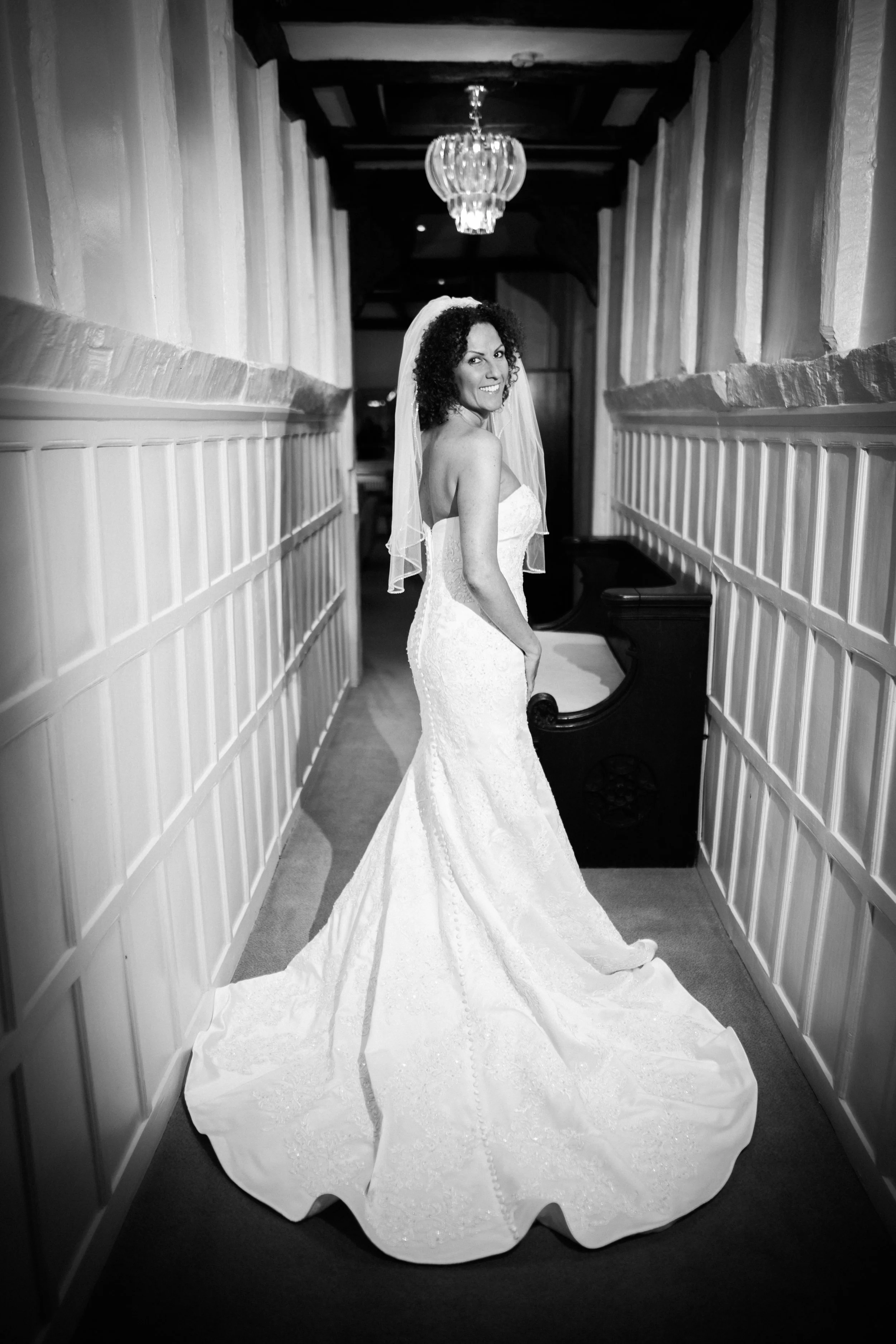 A bride in a wedding dress standing in a hallway, facing backward, with a veil and a chandelier overhead. Black and white photo.