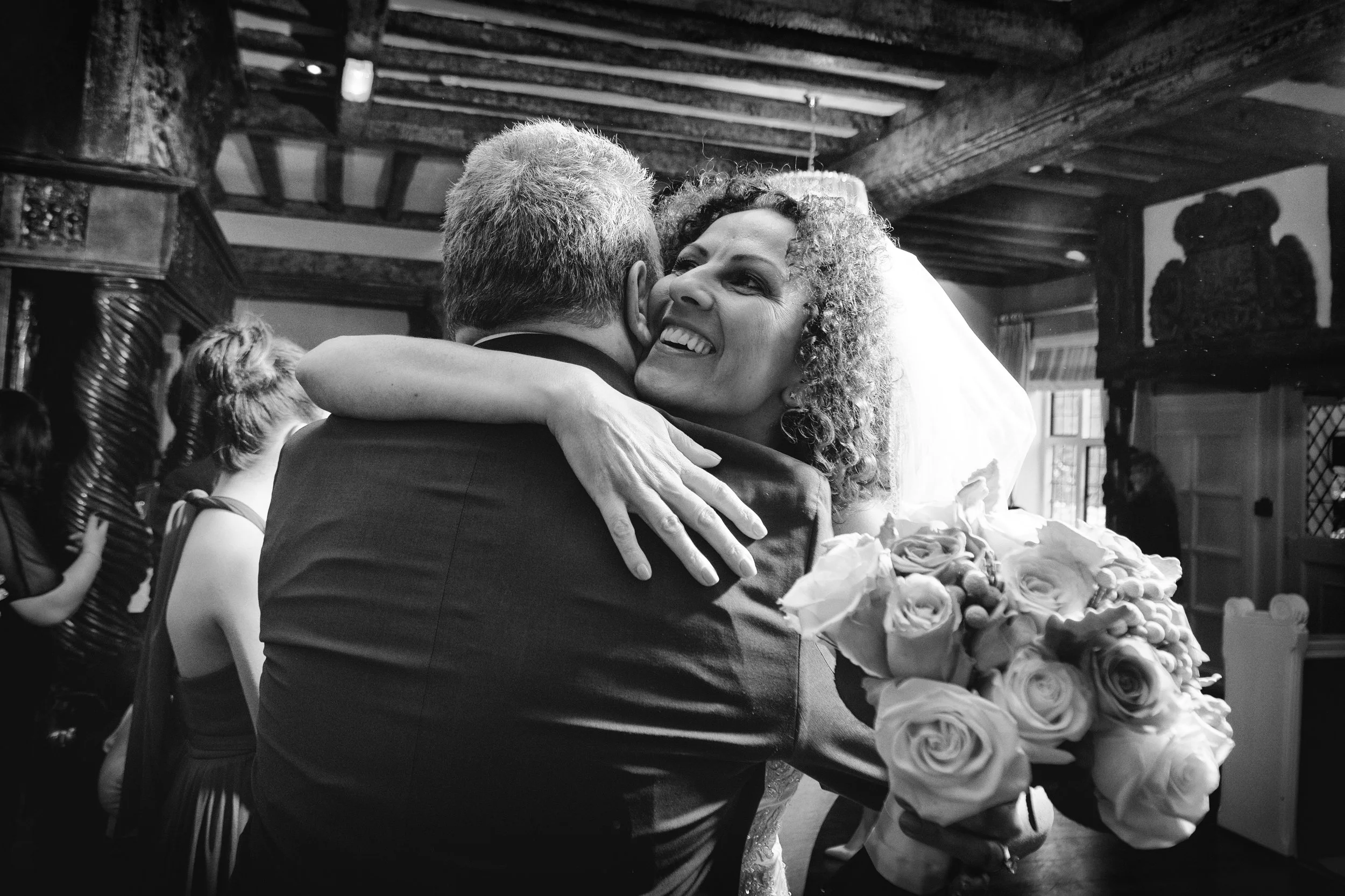 A happy bride embraces a person in a suit, holding a bouquet of roses in a warmly lit, rustic room.