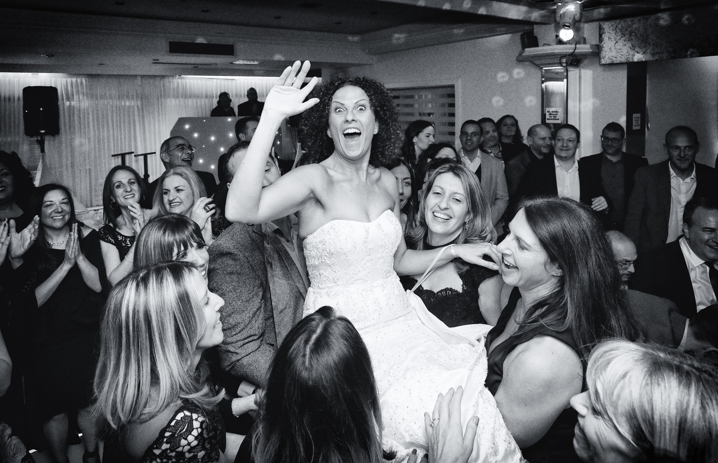Bride being lifted and celebrated by guests at a wedding reception.