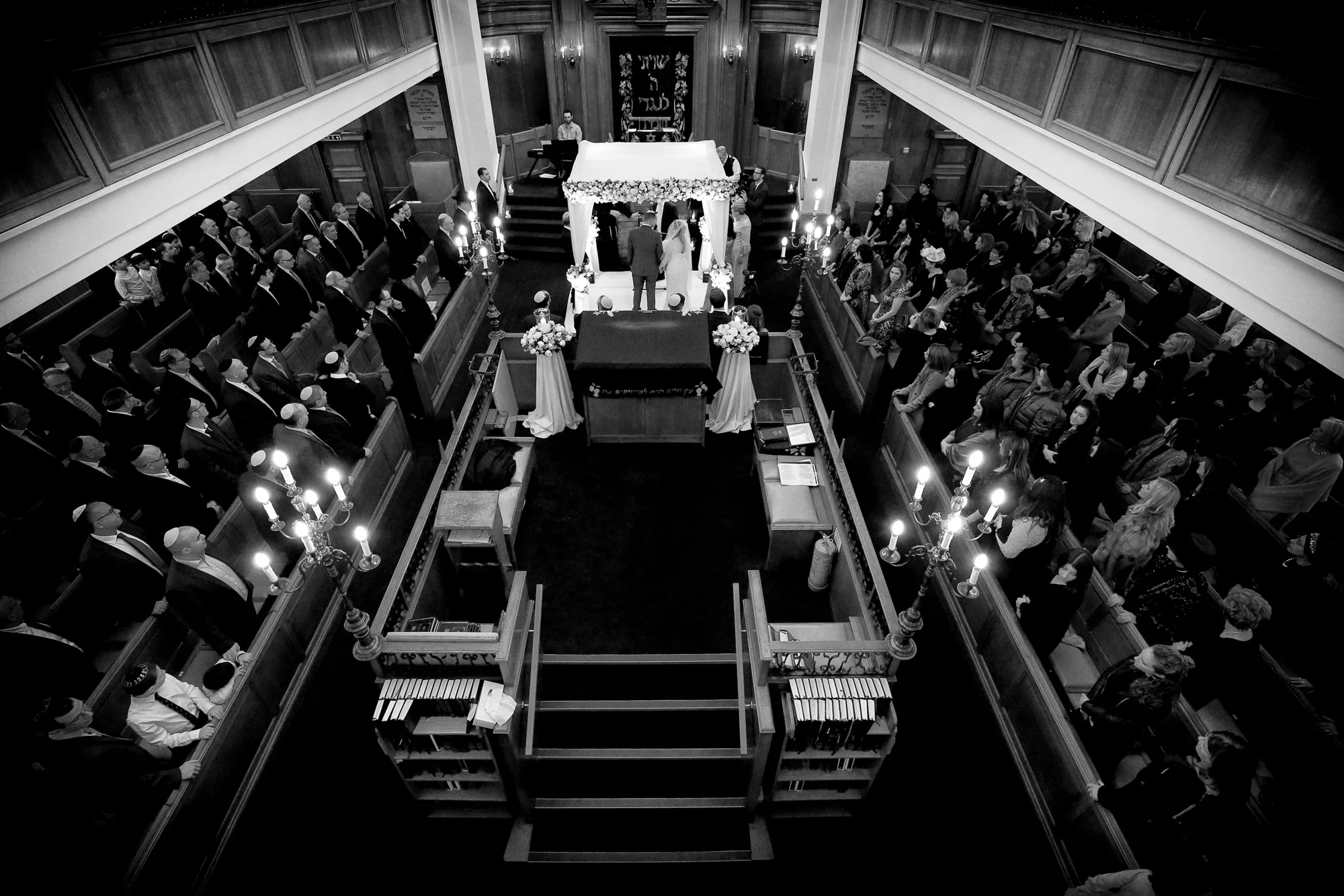 Black and white photo of a Jewish wedding ceremony in a synagogue, showing a bride and groom under a chuppah, surrounded by guests seated in wooden pews.