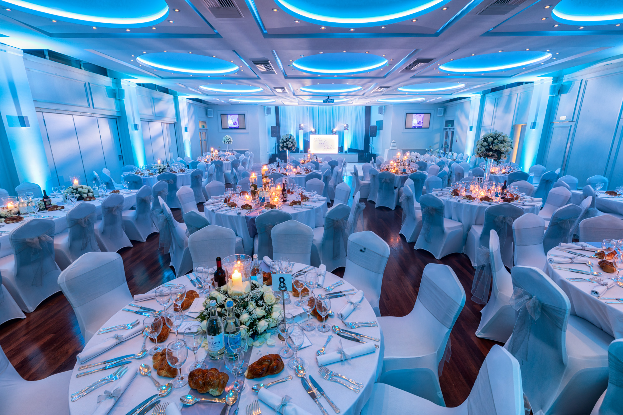 Elegant banquet hall with round tables set for an event, featuring white chair covers, floral centerpieces, candles, and ambient blue lighting.