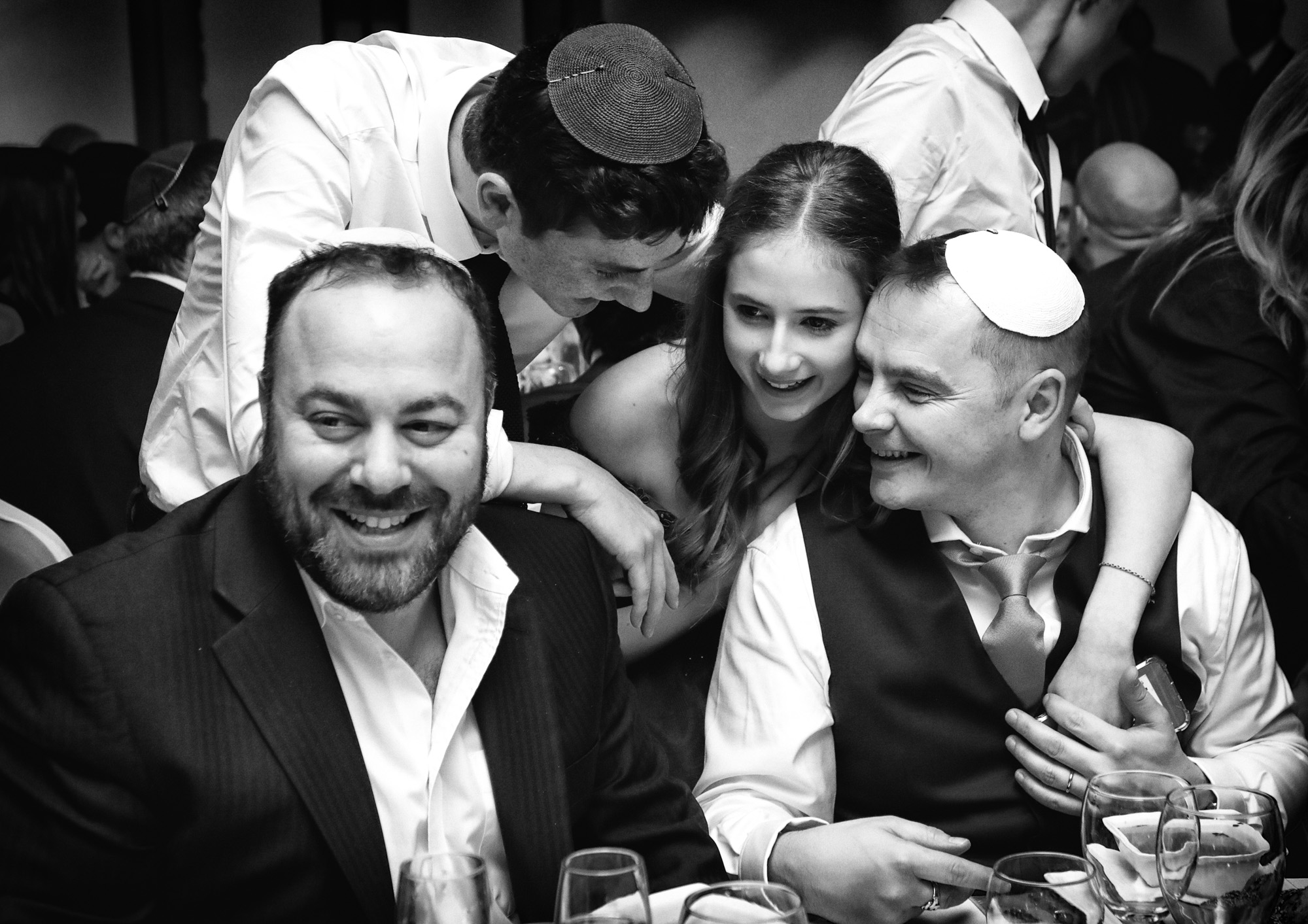 Black and white photo of people smiling, wearing formal attire, some with kippahs, at an event table.