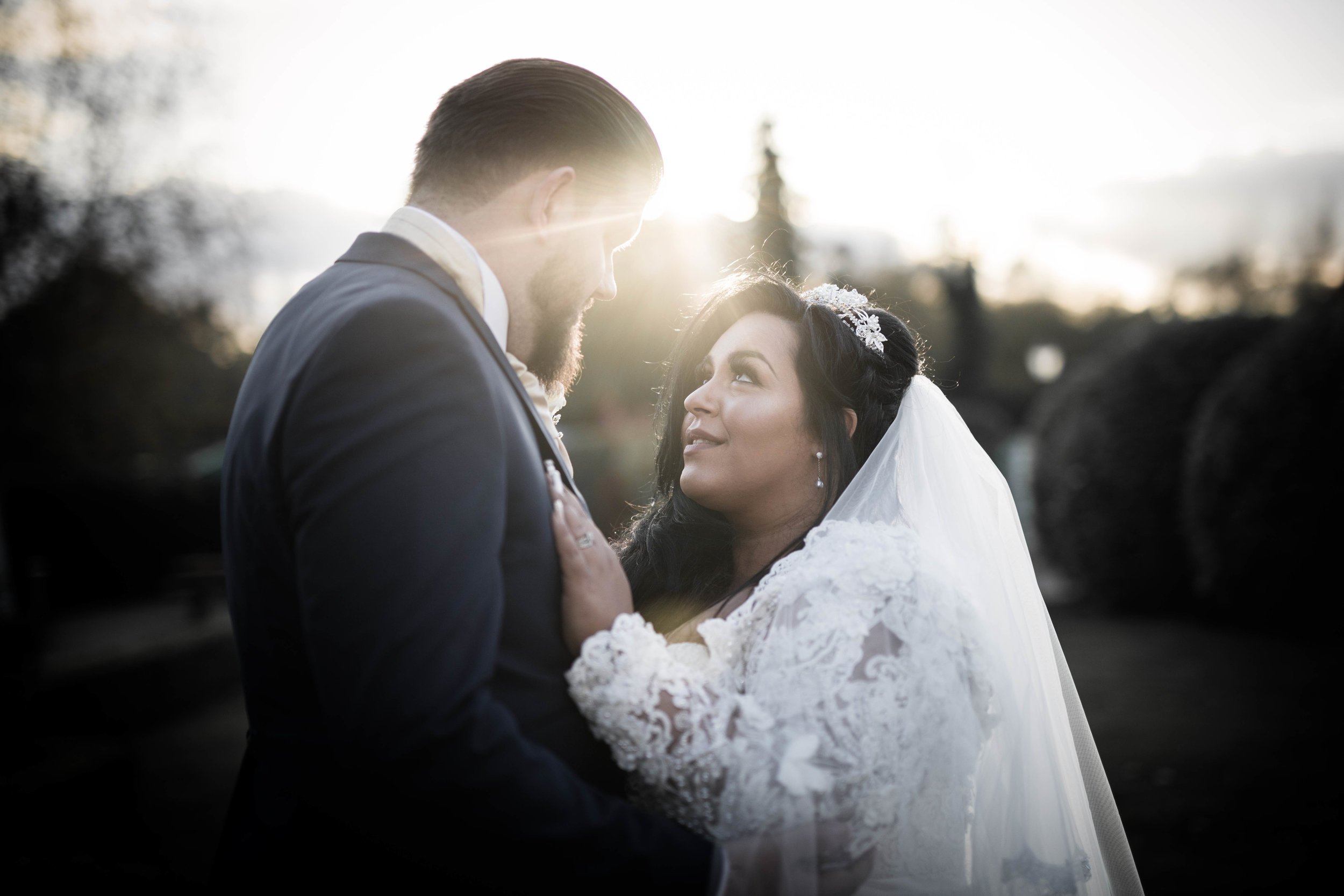 Bride and groom gazing at each other during sunset, with the bride in a lace wedding dress and veil, and the groom in a dark suit.