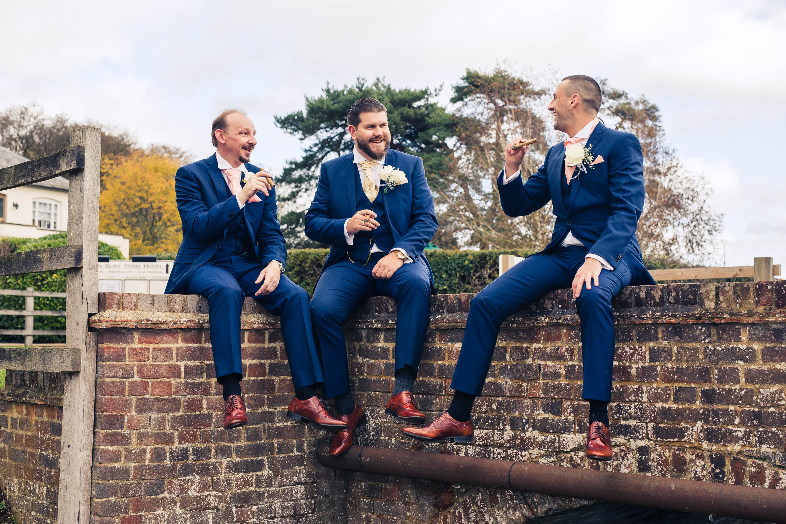 Three men in blue suits sitting on a brick fence, wearing brown shoes, holding cigars, and smiling outdoors.