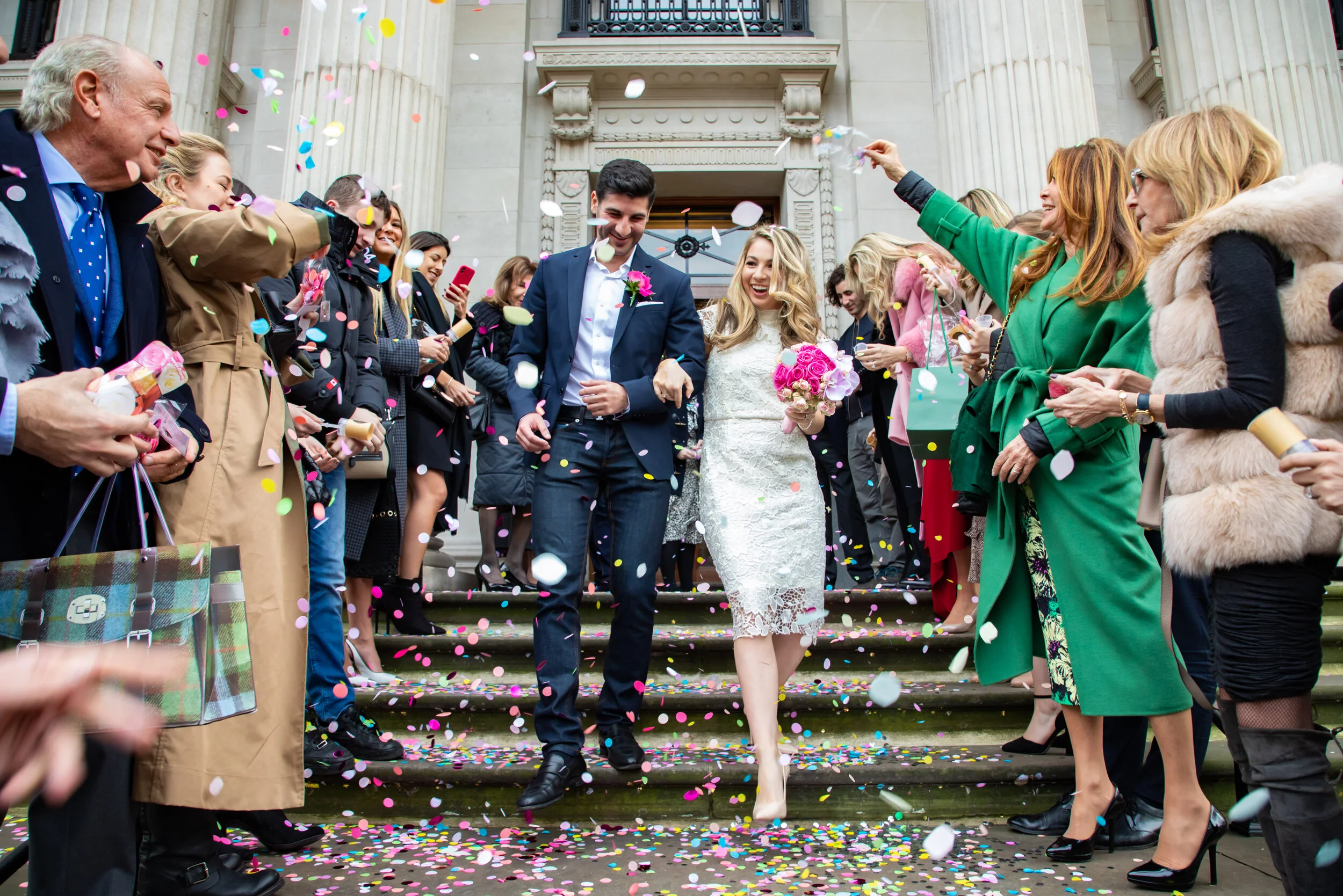 Bride and groom walking down steps surrounded by guests throwing confetti, at a wedding ceremony.