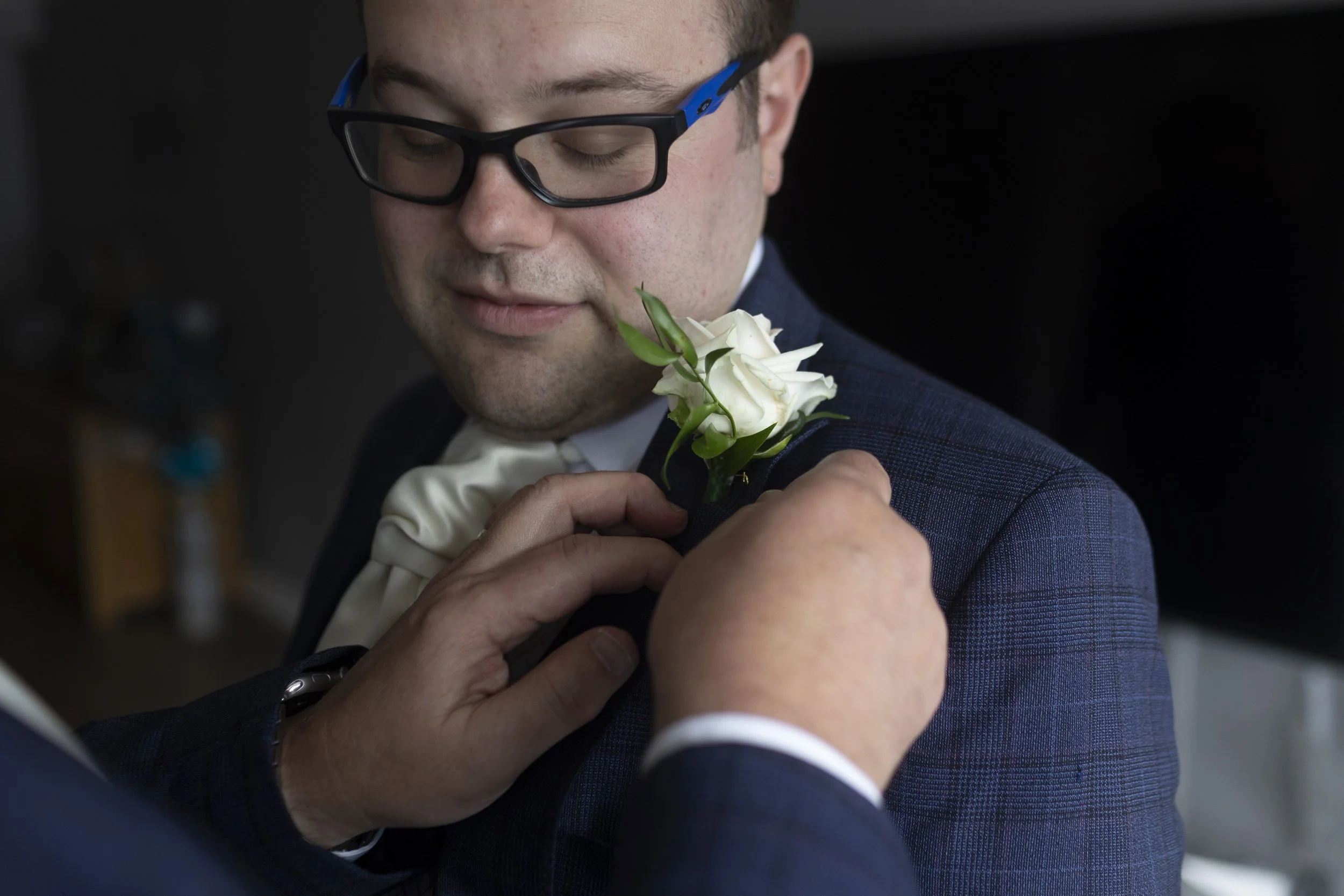 Man in glasses wearing a suit with a white rose boutonniere being adjusted by another person.