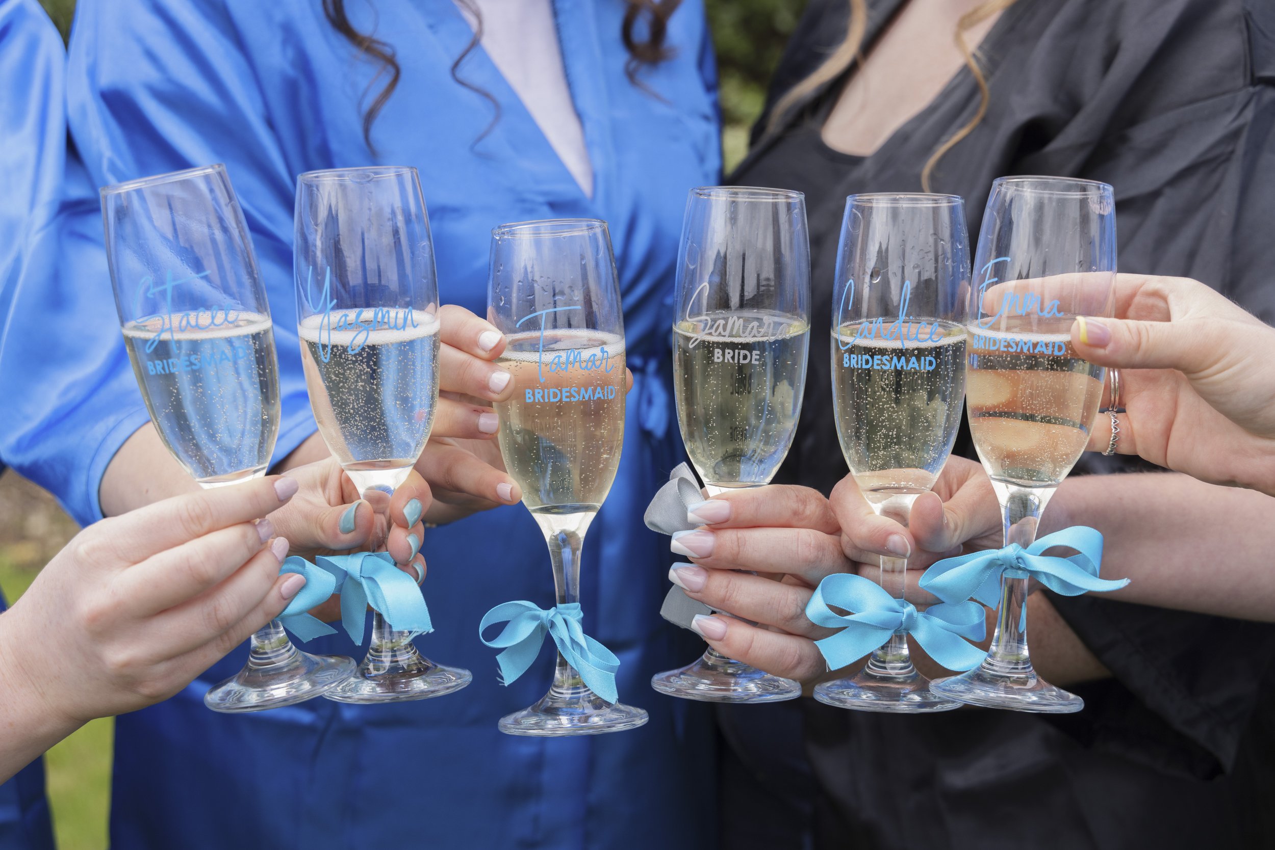 Group of women holding personalized champagne glasses with blue and gray ribbons, dressed in robes at a bridal party.