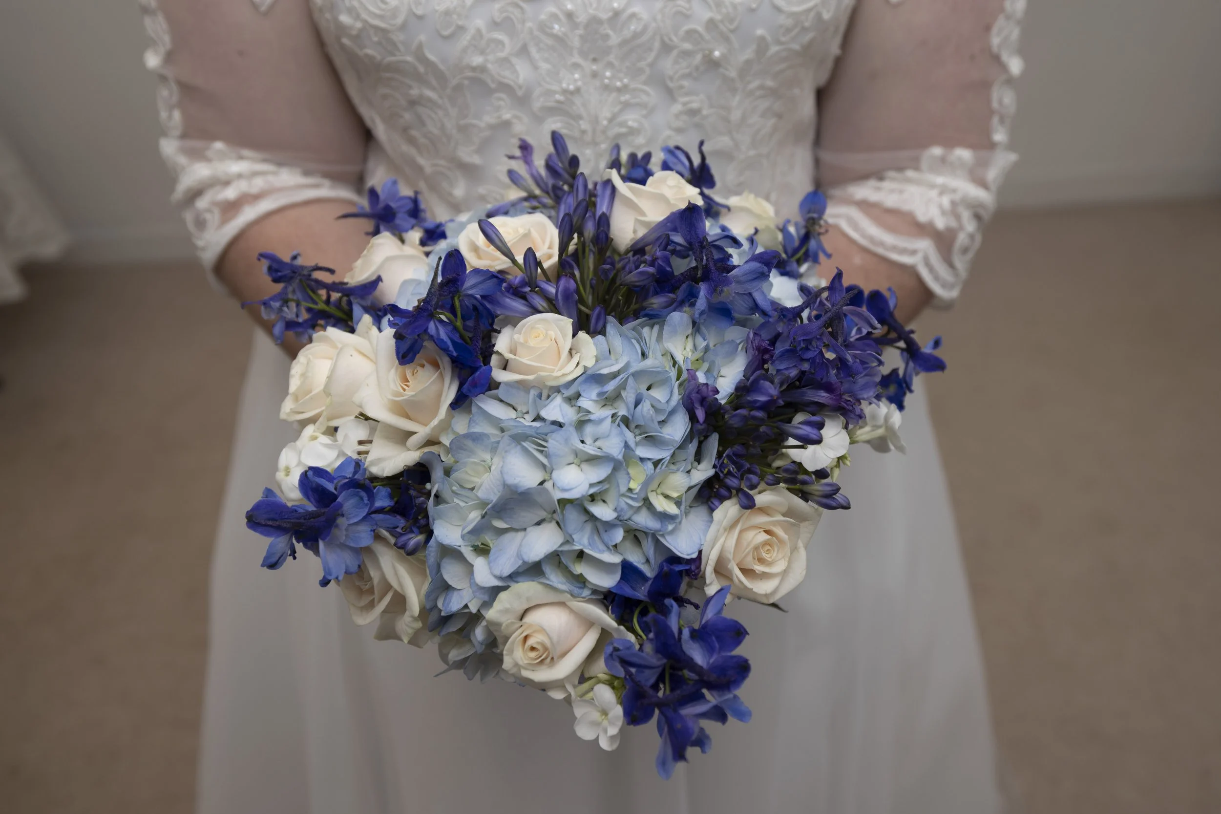 Bride holding a bouquet with white roses, blue hydrangeas, and purple delphiniums, wearing a lace wedding dress.