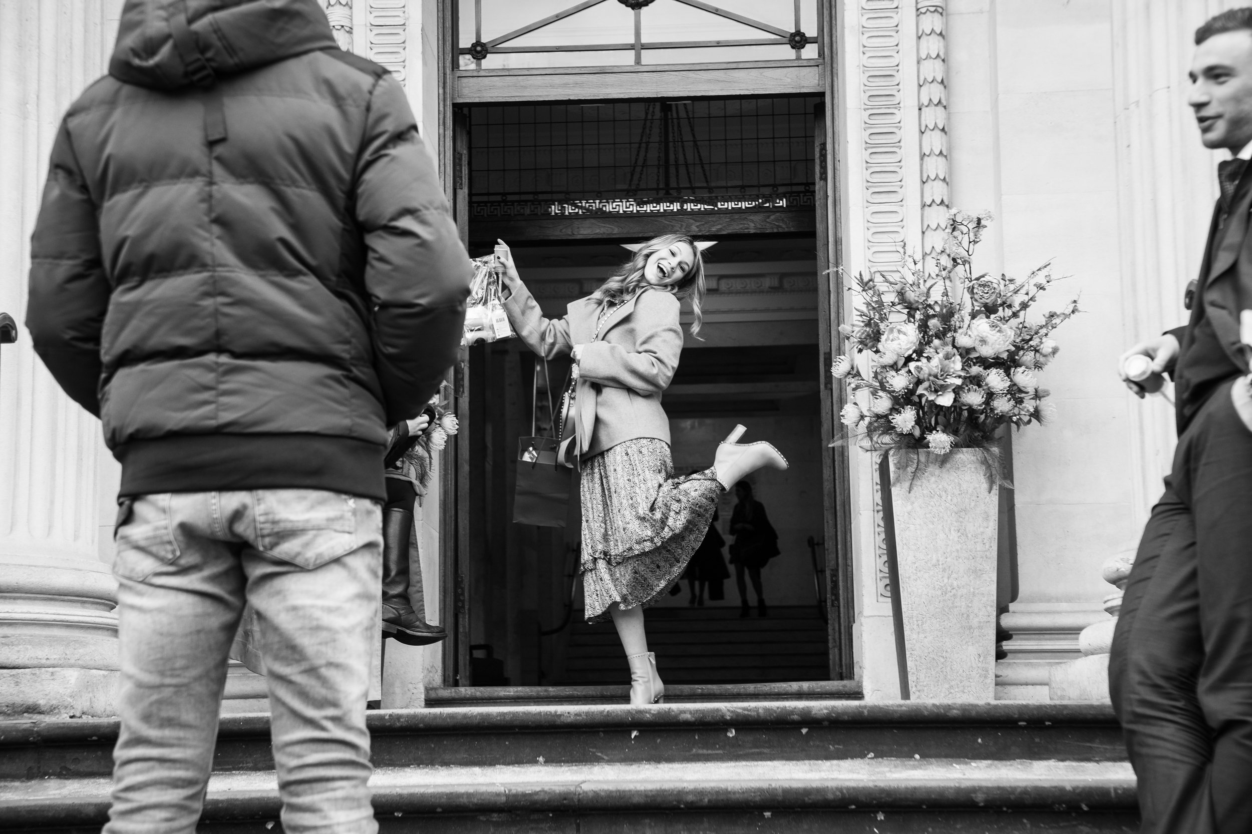 Black and white photo of a woman joyfully exiting a building, holding shopping bags, surrounded by other people and a floral arrangement on the steps.