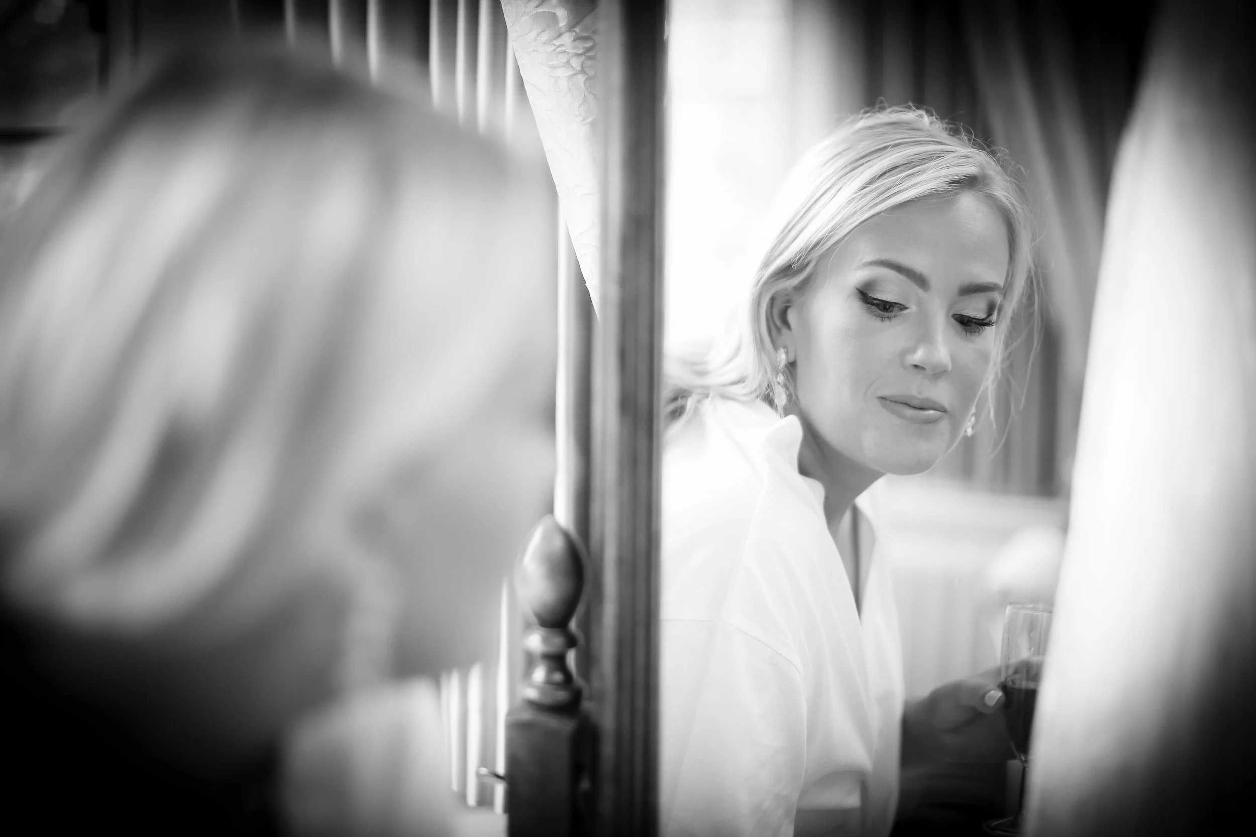 Black and white image of a woman with long hair and earrings, looking downward, seated near a curtain, holding a glass.